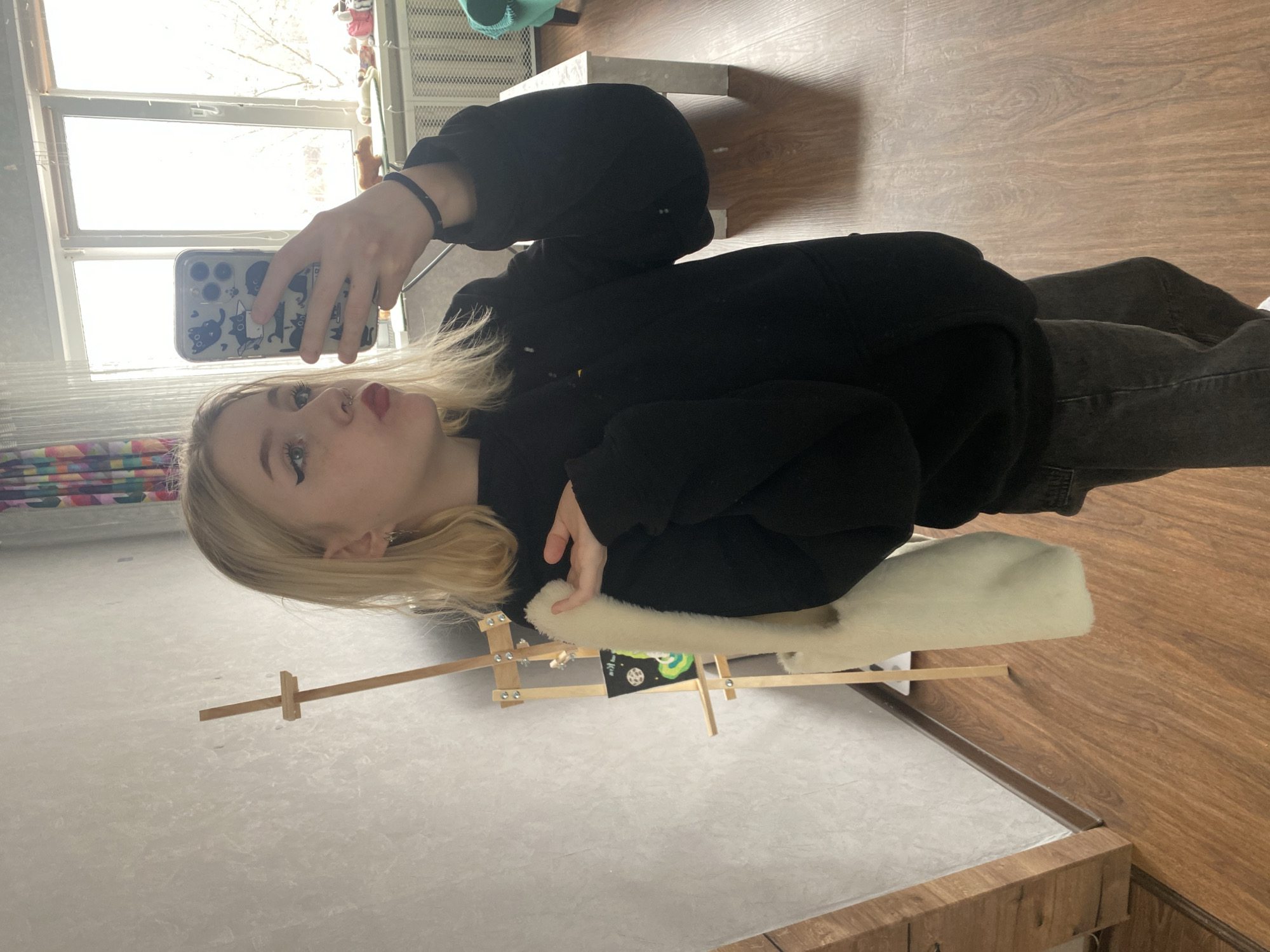 A young woman with long, light blonde hair and a nose piercing is taking a selfie indoors. She is wearing a black hooded sweatshirt and dark gray jeans. She holds a smartphone with a cat-themed case in her right hand. Her left hand is positioned near a small white pillow on an artist's easel. The easel holds a miniature painting featuring cosmic motifs. The background features a window with a view of an urban landscape, likely in Izhevsk, Russia, based on the room’s furnishings and architectural style. The room has a wooden floor and white walls. The lighting is bright and diffused.