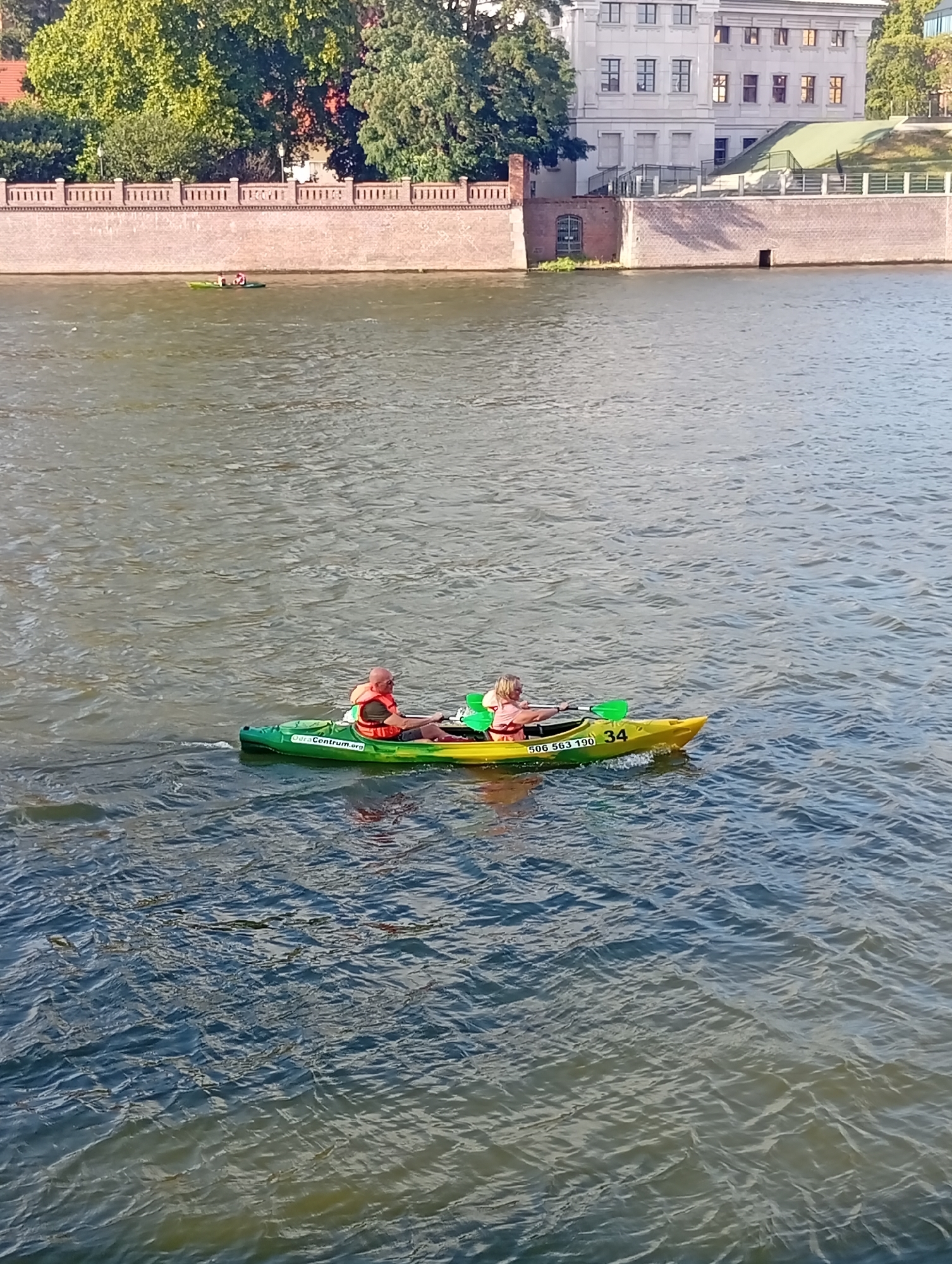 The image depicts a sunny day on the Oder River in Wrocław, Poland, with a view of the city's embankment and a building in the background. In the foreground, two individuals are kayaking. A man in an orange life vest and a bald head is in the rear of a green and yellow kayak, holding a paddle. A woman with blonde hair, also wearing an orange life vest, is in the front of the kayak, paddling with a green paddle. The kayak has markings that read "odraCentrum.org" and "506 563 190 34". The river water is choppy, reflecting the sunlight. The embankment features a brick wall with decorative elements and a lush green area beyond. The building in the background is a multi-story structure with many windows.