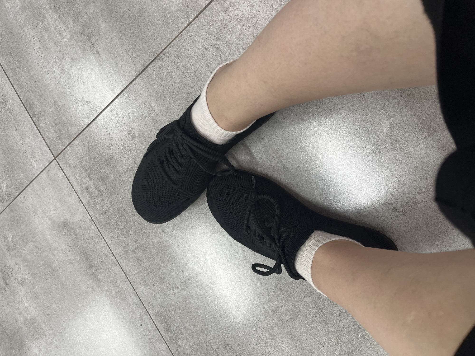 The image shows a person's legs wearing black sneakers and white socks, standing on a tiled floor. The scene is indoors, with the lighting suggesting it is daytime. The tiles are light gray with a concrete-like texture. The perspective is from a high angle, looking down at the legs and feet. The image does not provide any information about the city of Afipskiy, Russia, or any specific location within it. There are no other individuals or objects visible in the frame besides the person's lower legs, feet, shoes, socks, and the floor.