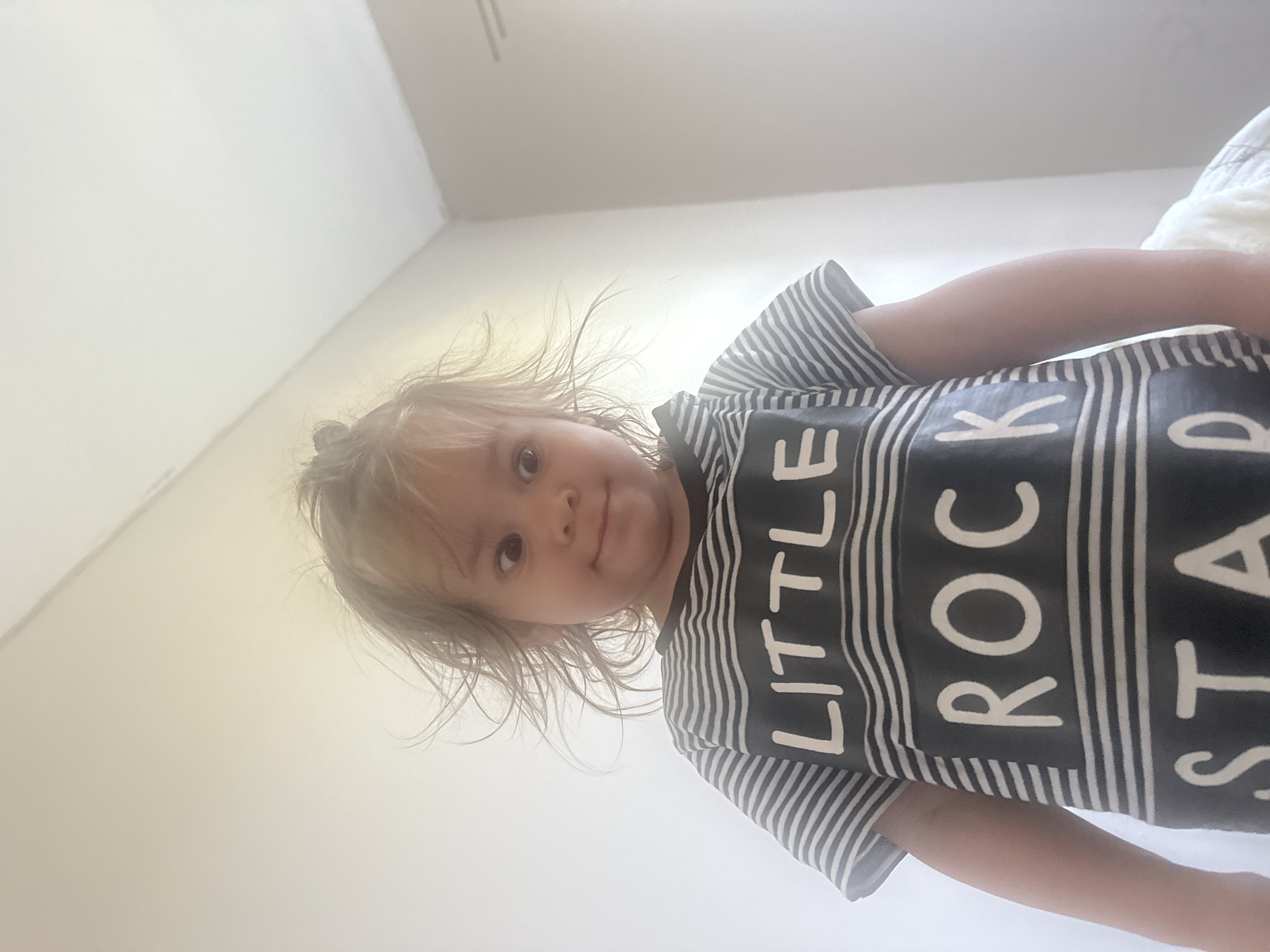 A young child with messy blonde hair stands facing the camera, their eyes wide and looking in slightly different directions. They are wearing a striped t-shirt that says "LITTLE ROCK STAR". The background is a plain, off-white wall, with a slight shadow in the upper right corner. The lighting suggests an indoor setting, likely a home. The image appears to have been taken from a low angle, looking up at the child. There is no visible indication of the city of Kharnavy, Russia.