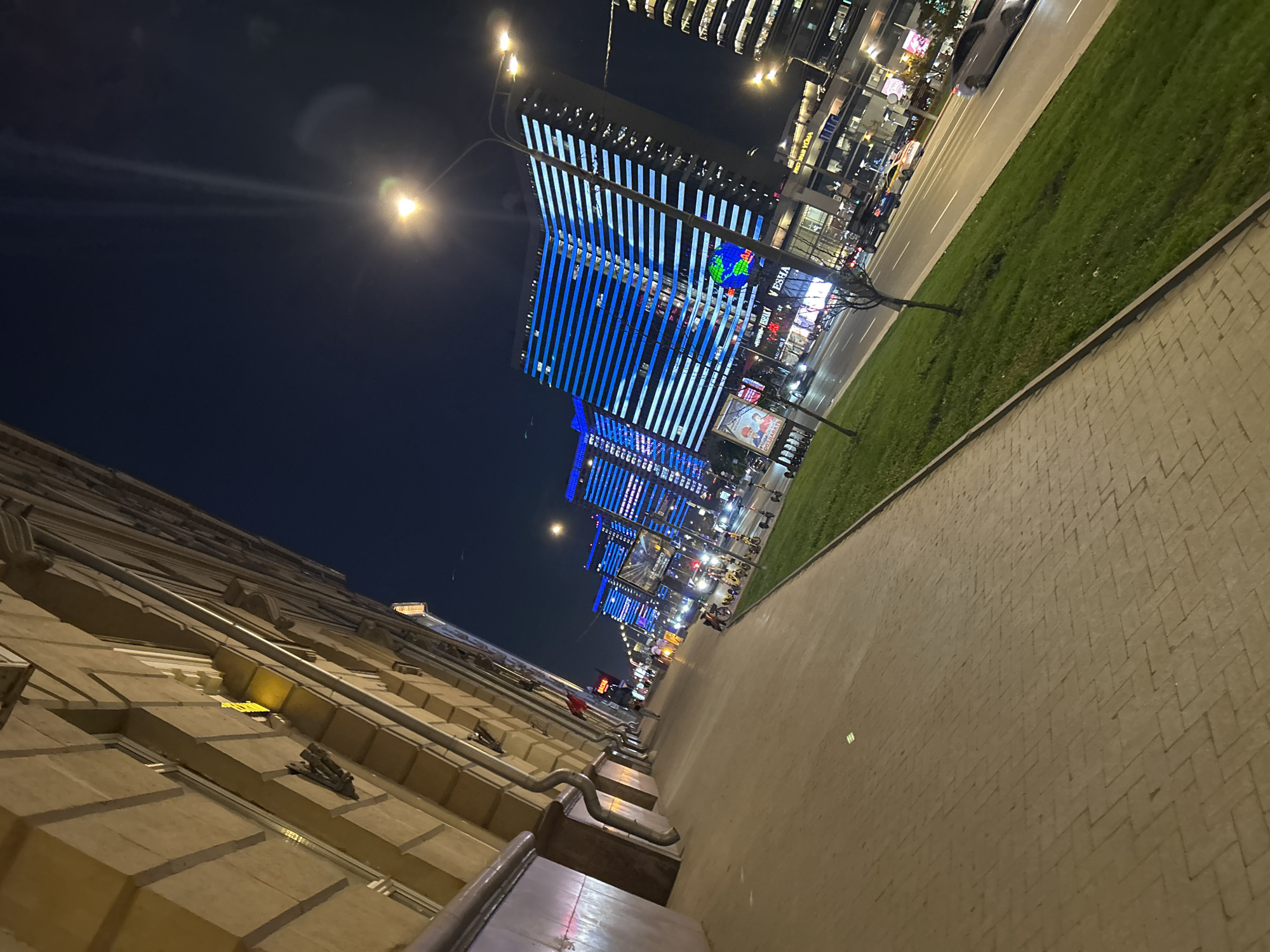 The image shows a street scene at night in an urban area, likely the city of Unknown, Unknown. The view is taken from a low angle, looking down a sidewalk towards a row of tall buildings. The buildings are illuminated with bright blue lights, arranged in vertical stripes. There are streetlights along the sidewalk, casting a yellow glow.
To the right of the sidewalk is a strip of green grass and a street with some cars. The sidewalk itself is paved with brick. The left side of the image shows the side of a building with a textured facade.