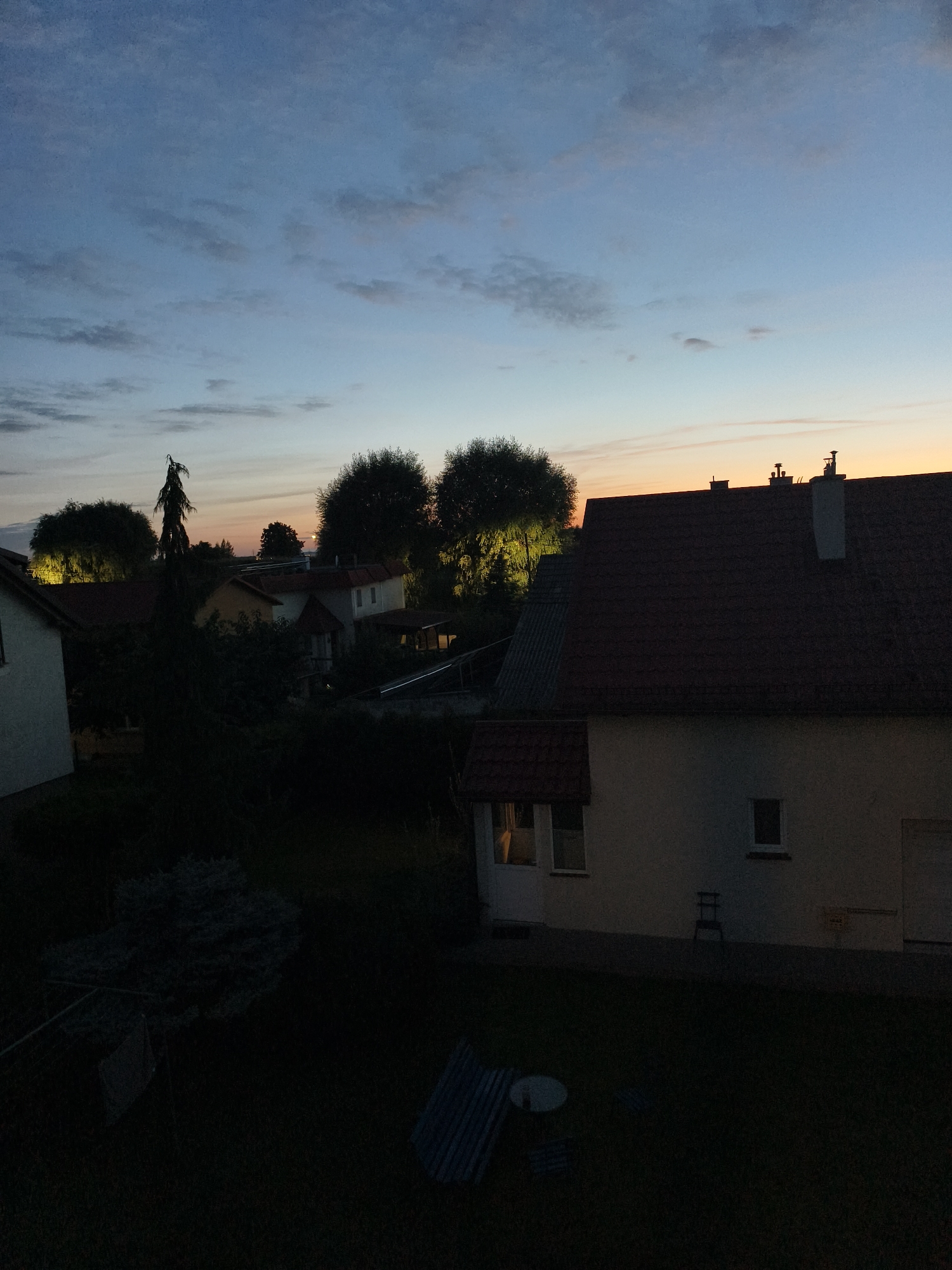 An exterior shot of residential houses and trees is captured at dusk or dawn. The sky is a gradient of blue, with scattered clouds transitioning to an orange hue near the horizon. Several houses with red-tiled roofs are visible, some with illuminated windows. Trees with dense foliage are interspersed throughout the scene. In the foreground, a dark lawn is visible with some garden furniture and debris. The overall impression is a quiet, suburban setting during twilight.