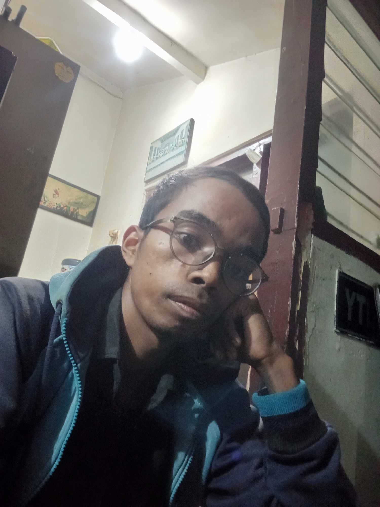 A close-up selfie shows a young man wearing glasses and a dark blue hooded jacket with a turquoise zipper and trim. He has his hand resting on his cheek, looking directly at the camera. The background appears to be an indoor setting with a light-colored wall, a wooden beam, and a framed picture on the wall. A fluorescent light fixture is visible on the ceiling. The overall scene suggests a casual, personal moment captured in what could be a home or office environment. The image does not contain specific details indicating the location within the city of Surabaya, Indonesia.