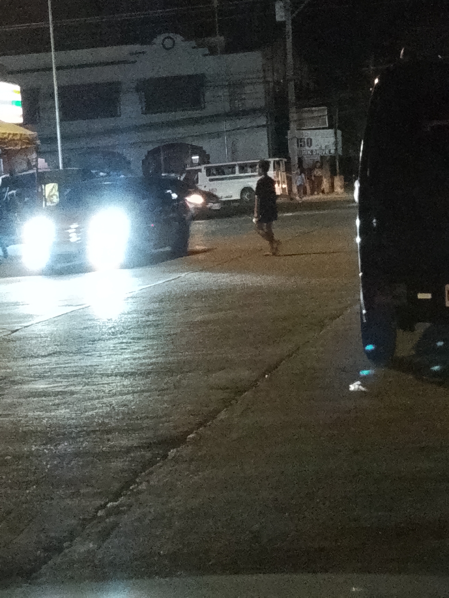 The image captures a nighttime street scene in Davao City, Philippines. The primary subjects are a person walking across a road and several vehicles, including a car with bright headlights illuminating the foreground. In the background, a building with dark windows and some signage is visible, along with other people standing near a bus. The lighting suggests it is nighttime, with artificial lights from vehicles and buildings being the main sources of illumination. The road appears to be paved and wet, hinting at recent rain or a damp environment.
The activity depicted is a person crossing the street, with vehicles present on the road. The presence of the bus and other vehicles suggests a public or semi-commercial area. The overall mood is that of a typical urban street at night, with the bright headlights creating a somewhat dramatic effect.
Notable details include the silhouette of the person walking, the intense brightness of the car's headlights, and the general urban setting. The building in the background has a circular decorative element above the windows. A sign with the number "150" and some illegible text is also visible in the background. The location context of Davao City, Philippines, places the scene within a specific urban environment in Southeast Asia. There are no clear indications of weather conditions other than the possibility of dampness on the road. The emotions conveyed are neutral, focusing on the visual aspects of the scene.