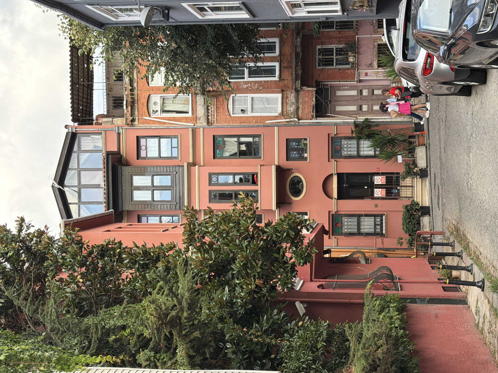This image depicts a narrow, cobblestone street in the Üsküdar district of Istanbul, Turkey, lined with historic buildings. The architecture features a mix of colors and styles, with a prominent salmon-pink building on the left showcasing traditional Ottoman architectural elements such as bay windows and ornate window frames. Adjacent to it are older brick buildings with more weathered facades, some featuring metal awnings and multiple windows. A grey building is visible on the right side of the frame. Lush green trees and vines cascade down the sides of some buildings, adding a touch of nature to the urban setting. Two women are standing and interacting in the middle of the street, with their backs to the camera. Several cars are parked along the street, with the rear of a silver sedan and a dark-colored vehicle in the foreground. The sky is overcast, suggesting a cloudy day.