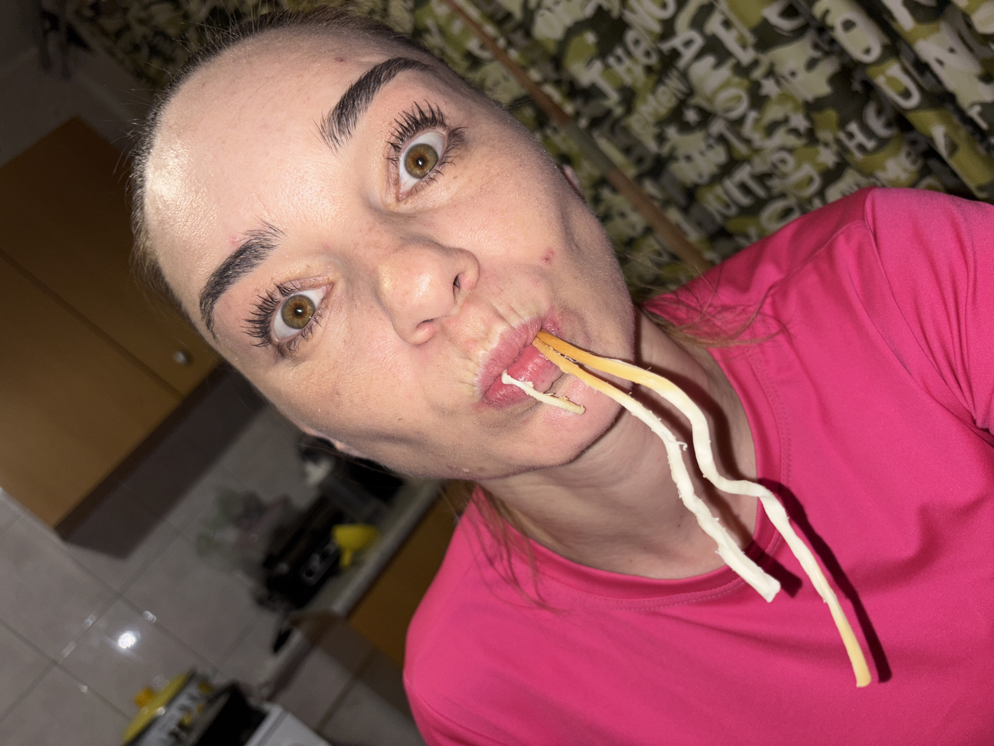 A woman, who appears to be in her kitchen, is captured in a close-up, head-and-shoulders shot. She is wearing a bright pink t-shirt. The woman has her mouth open, with several strands of what appear to be uncooked spaghetti noodles protruding from her mouth. Her eyes are wide open, and she is looking directly at the camera. The background shows kitchen cabinetry, tiled walls, and a patterned curtain, suggesting an indoor setting. The lighting appears to be artificial, possibly from overhead kitchen lights. There is no visible indication of her location in the city of Sinyavino, Russia.