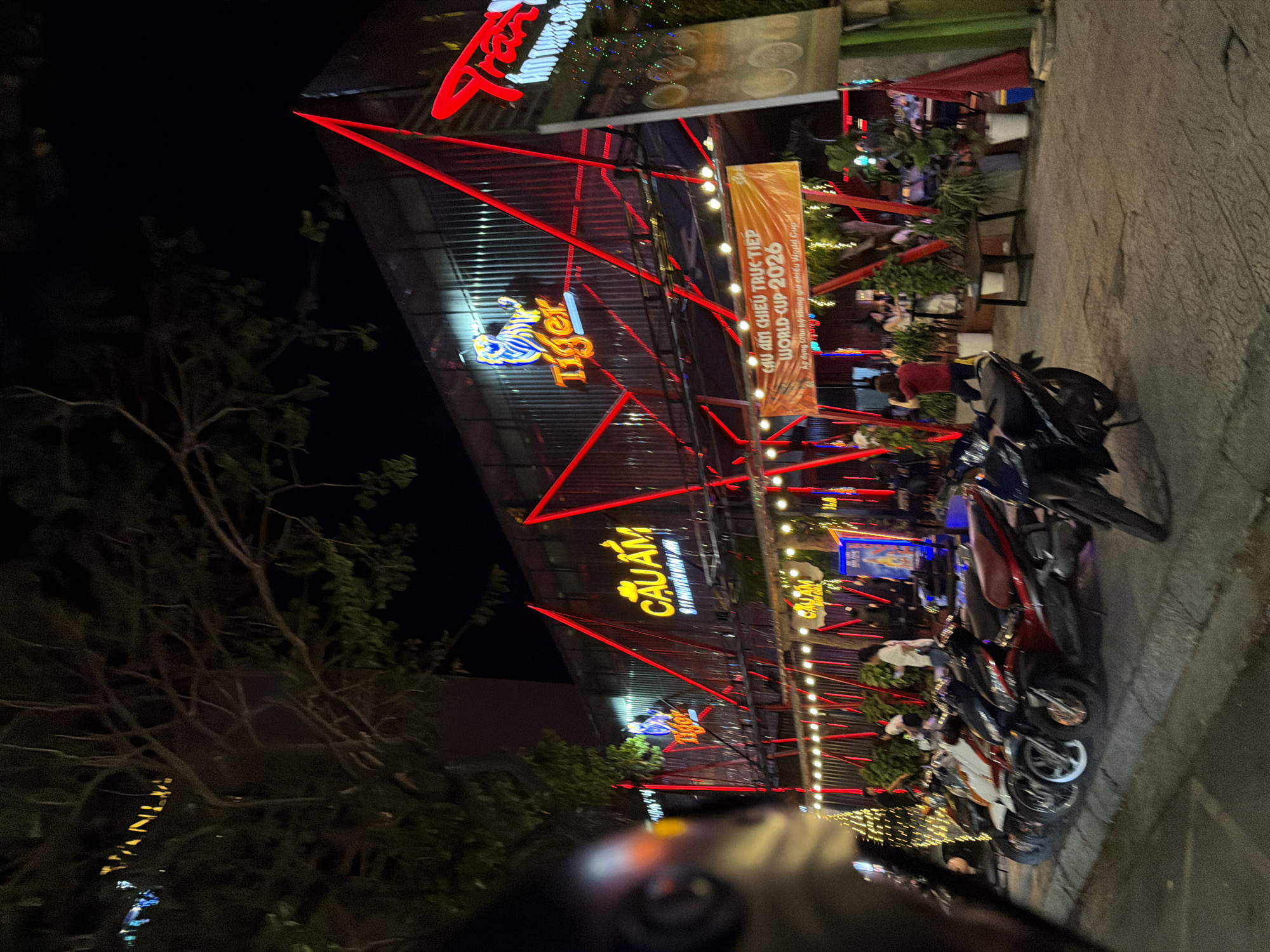This image captures a vibrant night scene at an outdoor eatery, likely in Đà Nẵng, Vietnam, judging by the text. The setting is illuminated by a profusion of neon lights and string lights, creating a festive and energetic atmosphere. Red neon tubes form an angular framework over the establishment, drawing the eye to illuminated signs.
Prominently displayed are signs for "Tiger," a well-known beer brand, and "CẦU AM," which appears to be the name of the eatery. One sign, in Vietnamese, announces "CẦU AM CHIEU TRUC TIEP WORLD CUP-2026," indicating a live broadcast of the World Cup in 2026. The presence of multiple motorcycles parked on the side suggests it's a popular spot for locals and visitors alike.
Several people are visible, seated at tables under the illuminated canopy, engaged in dining and socializing. The overall mood appears lively and social, typical of an evening gathering at a restaurant or bar. The time of day is clearly night, with the darkness of the background emphasizing the bright artificial lights. The weather seems pleasant, as people are dining outdoors. The composition, with the camera angled upwards and slightly skewed, adds a dynamic perspective to the scene.