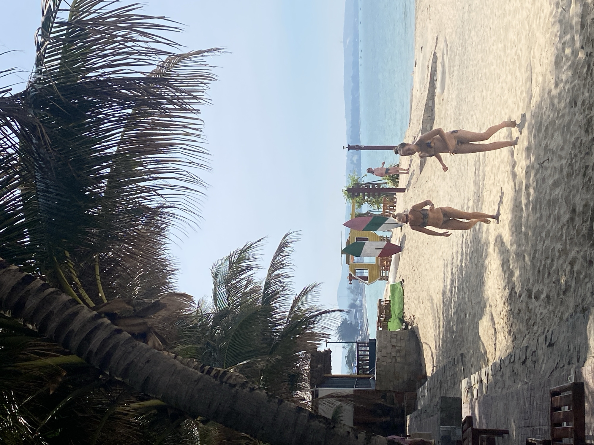 An outdoor, eye-level, medium shot captures a sunny day on a sandy beach in Phu Thuy Ward, Vietnam. The scene is dominated by a large palm tree in the foreground, its trunk a rough, textured grey and its fronds creating a dappled shadow across the sand. To the left, a rustic beachside building with a thatched roof and wooden railings is partially visible. The sandy beach extends towards the turquoise ocean, where several small boats are scattered on the horizon. In the middle ground, a yellow and white surfboard stands upright, next to a colorful surf-themed structure resembling a small shack or changing room. Two young women in bikinis are walking on the sand, one on the left and another on the right, both appearing to be in motion. Further in the background, a few other people are visible, including a woman standing near a wooden swing set and another person in the distance near the water. The sky is a clear, pale blue.