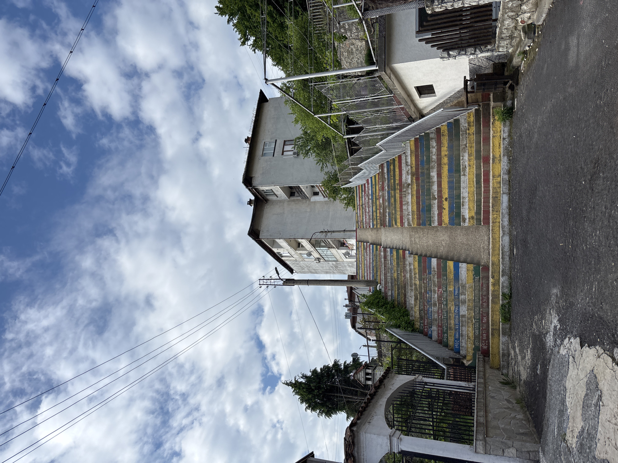 Here's a detailed description of the image:
The image shows a vibrant staircase in Zlatograd, Bulgaria. The stairs are painted with colorful horizontal stripes in blue, yellow, red, and green. Inscribed on the stairs are texts in Bulgarian, marking historical events.
To the right of the stairs is a narrow asphalt road, cracked and weathered. The stairs are bordered by metal railings.
Above the staircase, a building with a grey facade stands tall. Power lines are visible against the blue sky, punctuated by white clouds. An archway at the bottom of the stairs provides an entrance point from below.