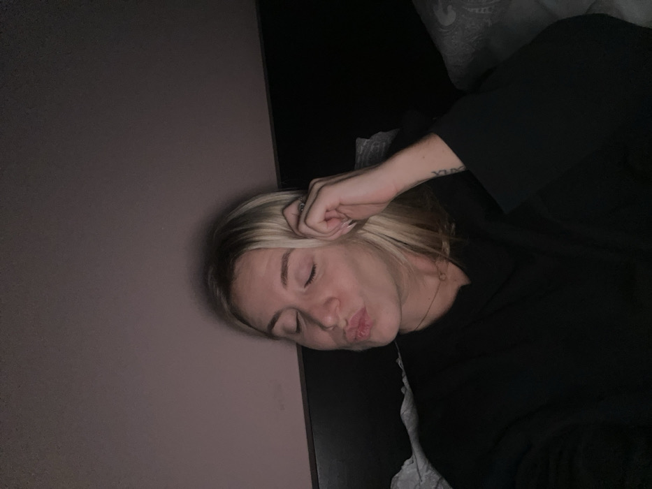 An adult female with blonde hair is depicted in a dimly lit indoor setting in Chambœuf, France. She is positioned slightly to the left of the frame, facing forward with her head tilted to the right. Her eyes are closed, and her lips are pursed in a kissing gesture. Her left hand is raised, with her fingers touching her left temple area. She wears a dark, long-sleeved garment, possibly a sweatshirt, and a thin necklace with a small, circular pendant is visible around her neck. A ring is present on a finger of her left hand, and what appears to be a tattoo, possibly Roman numerals, is visible on her inner left forearm. Behind her head and to her right, a dark, horizontal surface, possibly a headboard or backrest, is visible. To the right of the frame, soft, grey patterned fabric, likely bedding or a cushion, is present. The background above her head features a plain wall with a muted pinkish-purple hue, illuminated by soft, indirect light, creating a warm ambiance.