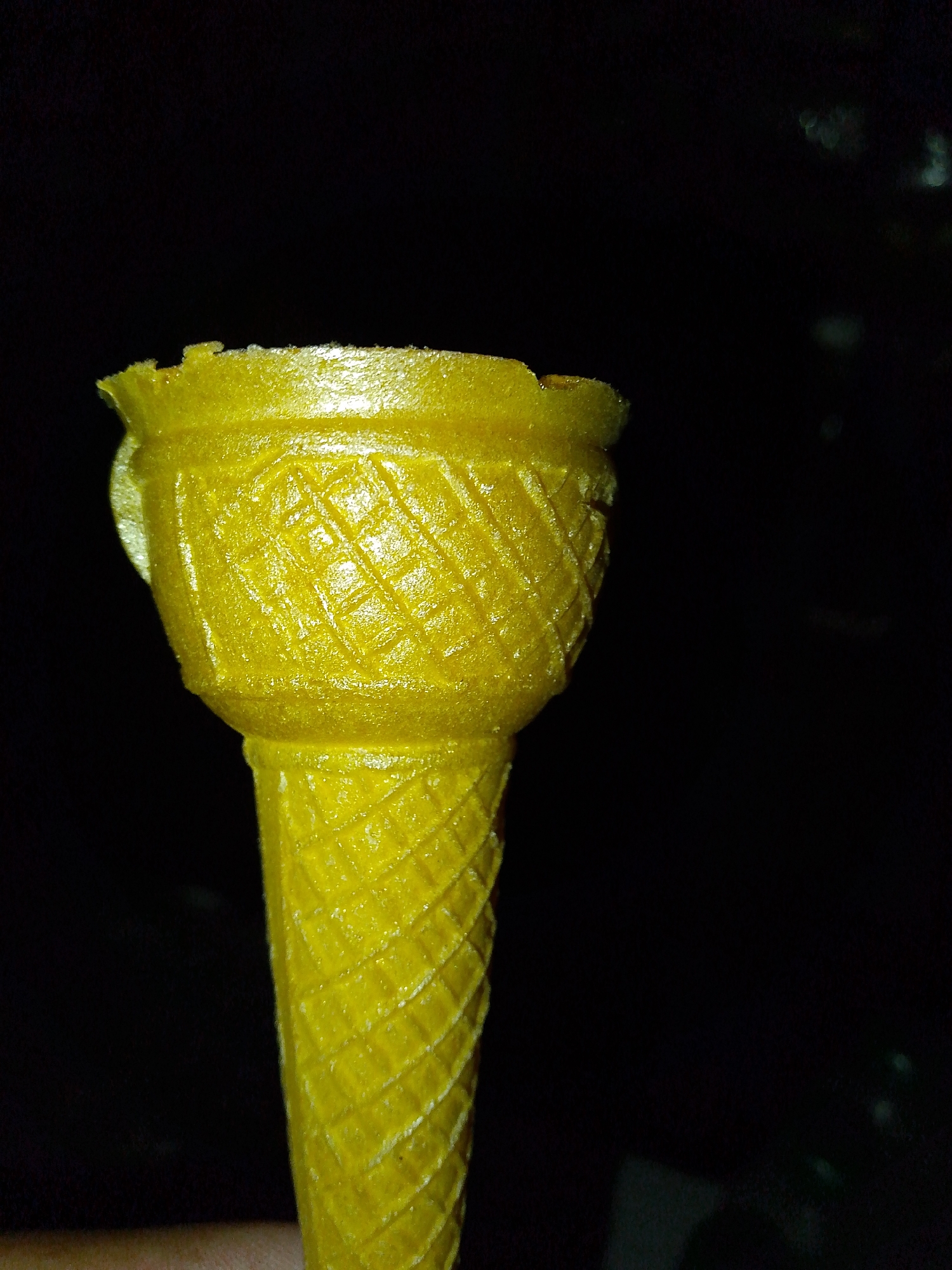 The media file is a close-up, vertical shot of a yellow ice cream cone against a dark, out-of-focus background. The cone is the primary subject and is brightly lit, showcasing its textured surface. The top portion of the cone has a flared rim with a decorative, diamond-like pattern. Below the rim, the cone tapers down, continuing the same patterned texture. The lighting highlights the glossy finish of the cone, with some areas appearing brighter than others, suggesting the direction of the light source. The background is predominantly black, with a few faint, out-of-focus blurry shapes that could be distant lights or objects. No people, text, or specific location cues are visible in the frame. The overall impression is a still life photograph focusing on the details of the ice cream cone. The dark background creates a strong contrast, making the yellow cone stand out prominently. The image appears to have been taken at night or in a dimly lit environment.