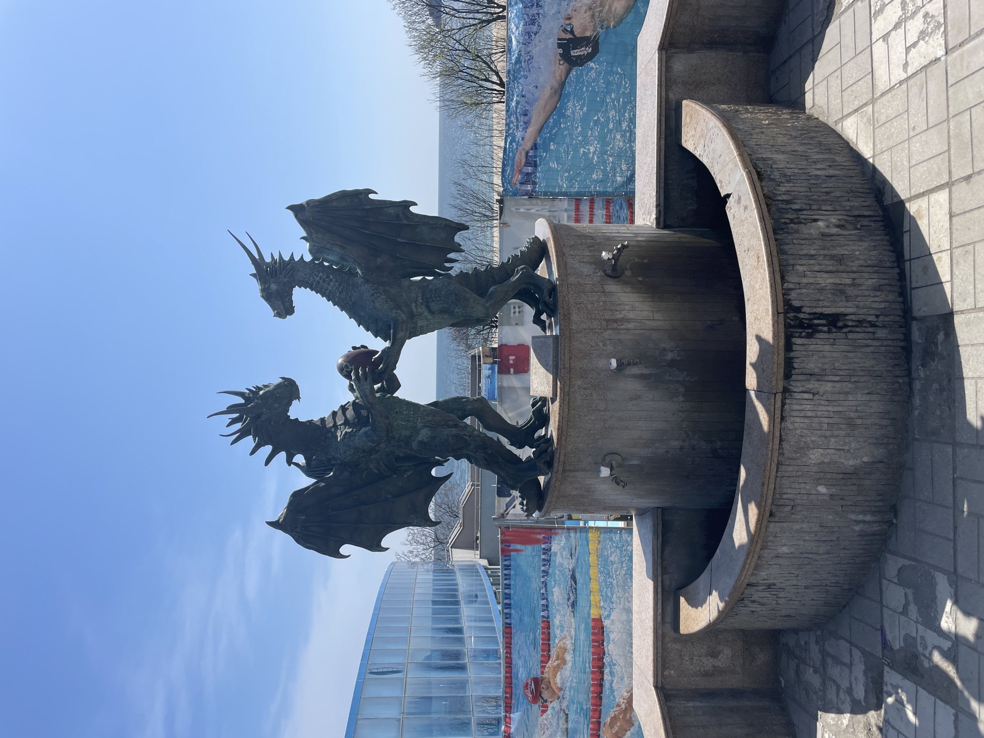 The image shows a statue of two dragons holding a red sphere, likely located in Varna, Bulgaria. The statue is mounted on a circular, tiered stone base. Behind the statue, there are outdoor advertisements featuring swimmers in a pool. A modern, glass-facaded building is visible to the left, and a glimpse of the sea and the horizon can be seen in the background. The sky is clear and blue. The image appears to have been taken during the daytime. The statue and its base are positioned on a paved area, and a portion of a curved stone structure is visible on the right.