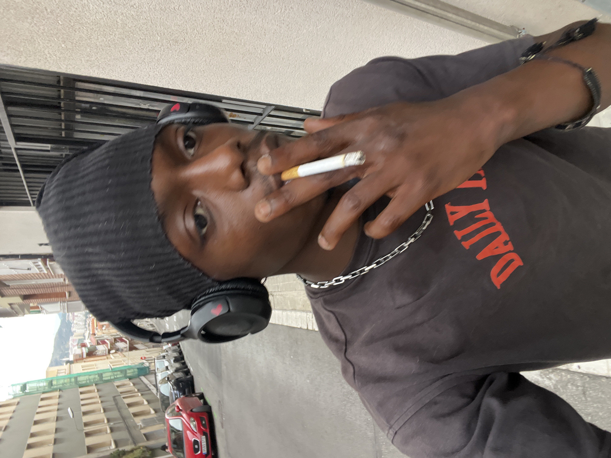 A man of African descent, wearing a black beanie and headphones, is smoking a cigarette. He has a silver chain necklace and a bracelet. The T-shirt he is wearing has "DAILY" written in red. The location appears to be a street in Barcelona, Spain, with buildings and cars visible in the background.