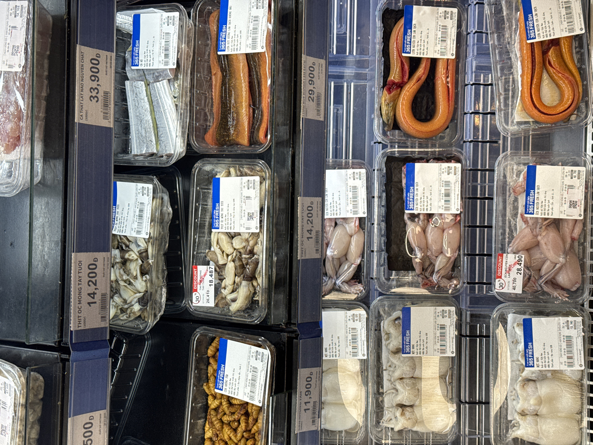 The image depicts a refrigerated display case in a grocery store, showcasing various packaged meats and seafood. The products include eel, fish, frogs, squid, and silkworm larvae. Each package has a label with the product name, weight, price in Vietnamese Dong (VND), and the "365 FRESH" brand. A "30% DISCOUNT" sticker is visible on some products. The store seems to be located in Vietnam, judging by the currency and Vietnamese text on the labels. The scene suggests a retail environment focused on selling fresh, packaged food items.