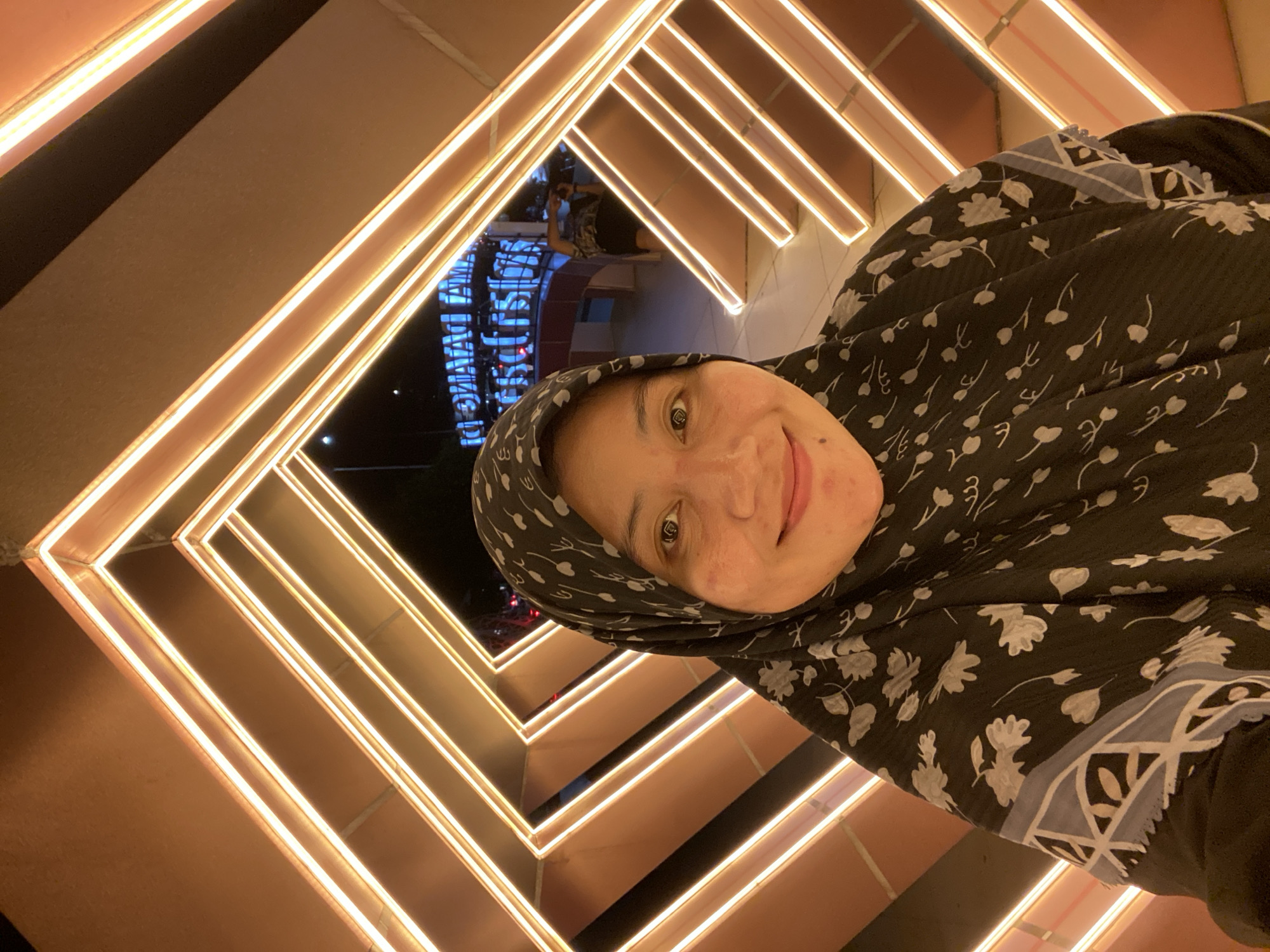 A woman wearing a black and white floral hijab and a black dress with floral and geometric patterns is taking a selfie. She is smiling at the camera. Behind her, there is an illuminated art installation with geometric shapes and orange neon lights. In the background, a blurry blue sign reads "DLAMANG AM" and "PARISIS" which likely refers to a location or event in Banjarbaru, Indonesia. There is another person in the background, blurred, holding a camera. The scene appears to be outdoors at night.