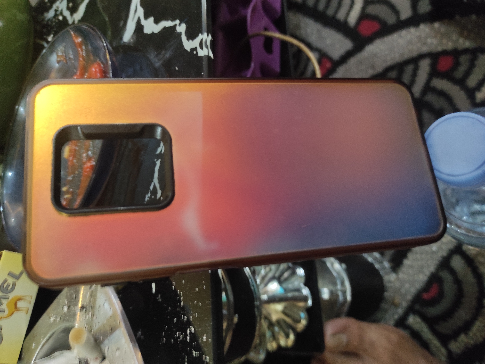 The image shows a smartphone case with a gradient color from orange to purple, resting on a patterned surface. The case has a cutout for the camera lens. In the background, there is a cigarette pack with a camel logo, an ashtray with cigarette butts, a metal object, and a bottle of water. The scene appears to be indoors, possibly on a table. The image is taken from a slightly elevated angle, focusing on the smartphone case. There are no people clearly visible in the image, though a foot can be seen in the lower left corner. The environment is cluttered with various objects, suggesting a casual or personal setting. The location in the city of Sumedang, Indonesia is not discernible from the visual information provided in the image.
