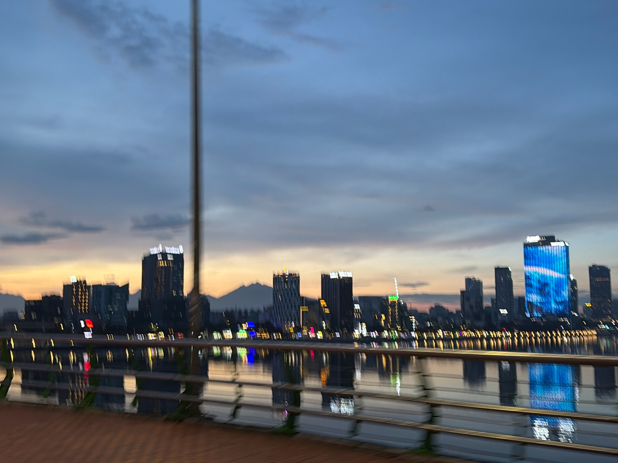 A blurred, motion-captured photograph depicts the cityscape of Đà Nẵng, Vietnam, at dusk. The sky is a blend of deep blues and soft oranges, with wispy clouds. A prominent lamppost stands vertically in the left half of the frame, its presence slightly obscuring the view.
The city skyline is characterized by numerous high-rise buildings, many of which are illuminated with colorful lights. One building on the right is particularly striking, featuring a large blue digital display that shows a dynamic, wave-like pattern. The lights of the buildings are reflected in the calm water of a river or bay that runs horizontally across the lower half of the image.
In the foreground, a metal railing with horizontal bars runs along the edge of a walkway or pier. The walkway itself appears to be paved with reddish-brown bricks or tiles, laid in a patterned manner. The overall impression is one of a vibrant, modern city viewed during twilight, with the movement of the camera adding a dynamic, somewhat abstract quality to the scene. There are no discernible people or specific actions captured in the photograph due to the motion blur. The image captures a section of the Han River waterfront.