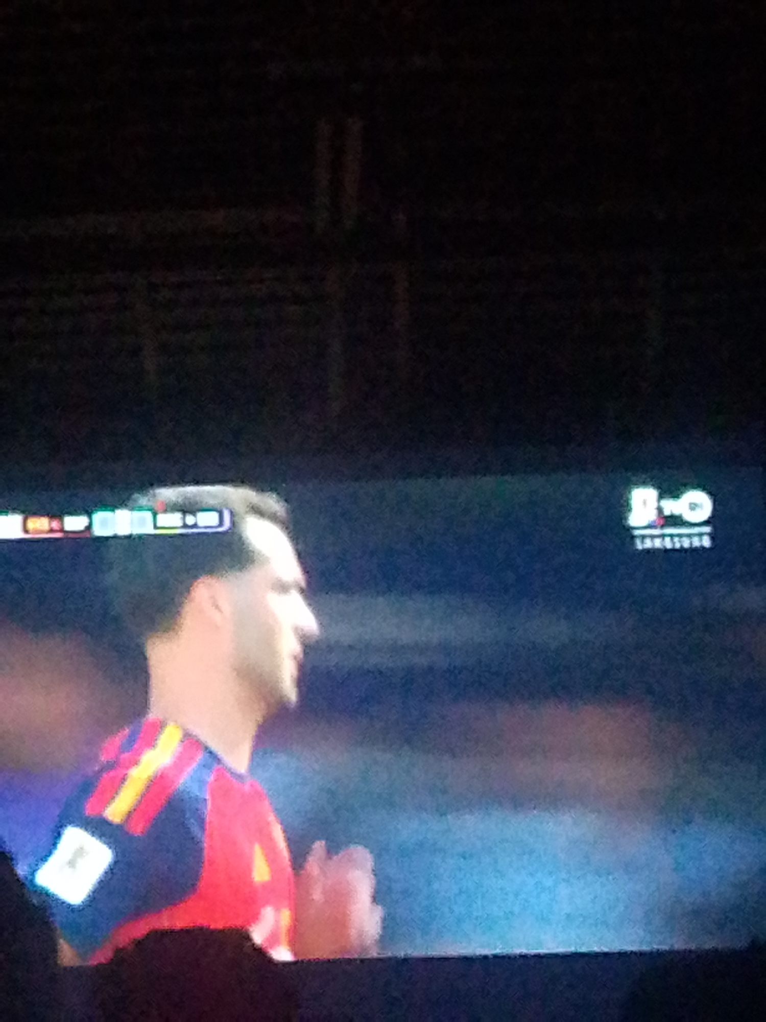 The image shows a close-up, profile view of a male soccer player on a television screen. He is wearing a red, blue, and yellow striped jersey. A white square patch is visible on his left shoulder. The player has dark hair and is looking to his right. A digital display bar with colorful squares is visible above his head, and a logo with the letters "D-N-O" and "LAKOTONG" is displayed on the right side of the screen. The background of the screen is a blurred blue and black. The image appears to be taken from a low angle, possibly in Meunasahmee, Indonesia, where the soccer match is being broadcast.