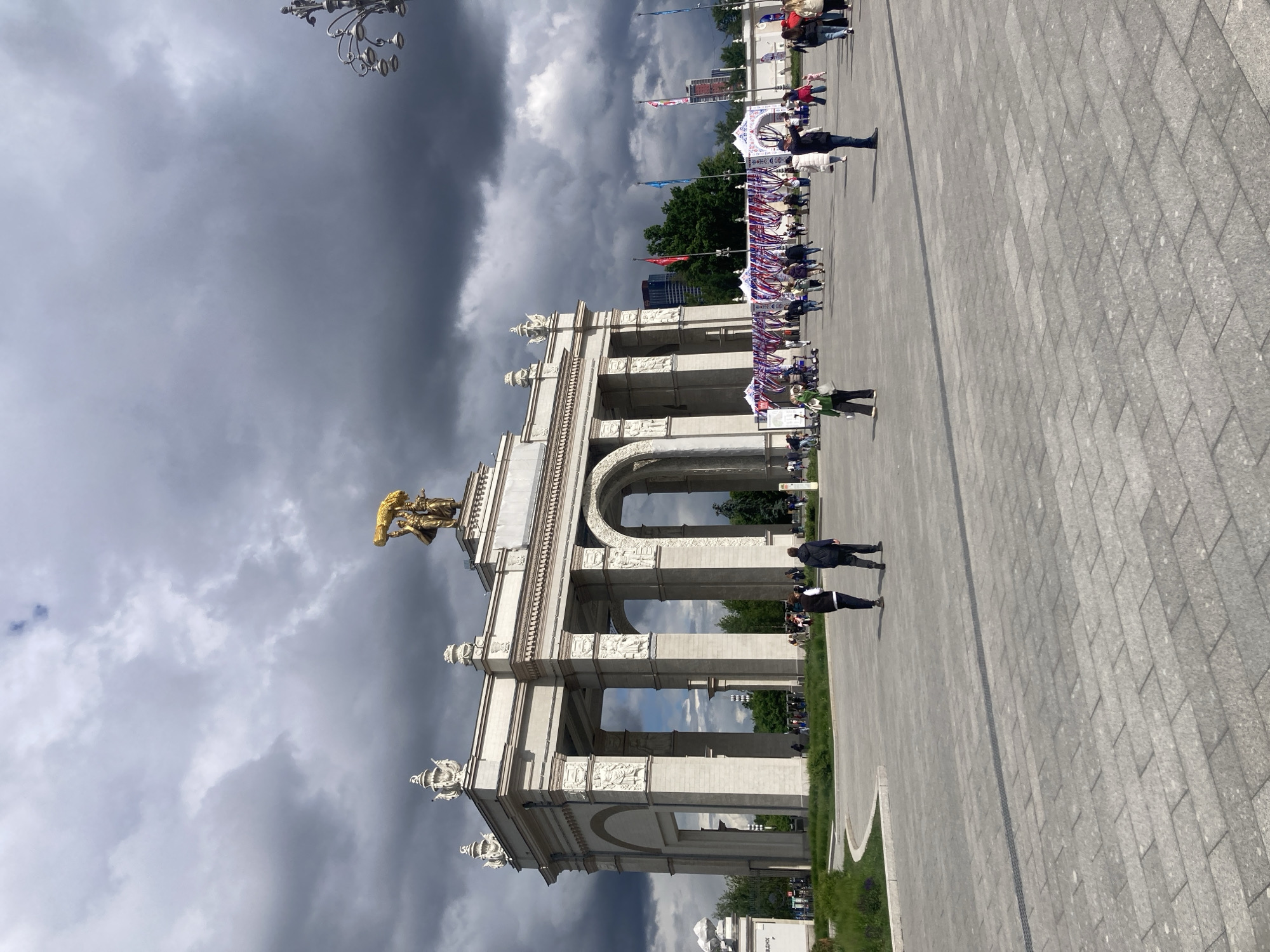The image shows a large, ornate, white architectural archway with a golden statue atop. The sky is overcast with dark, ominous clouds. People are walking on a wide, paved walkway in front of the archway. Flags and banners are visible strung along the structure. This is most likely the Central Pavilion at the Exhibition of Achievements of National Economy (VDNKh) in Khimki, Russia.