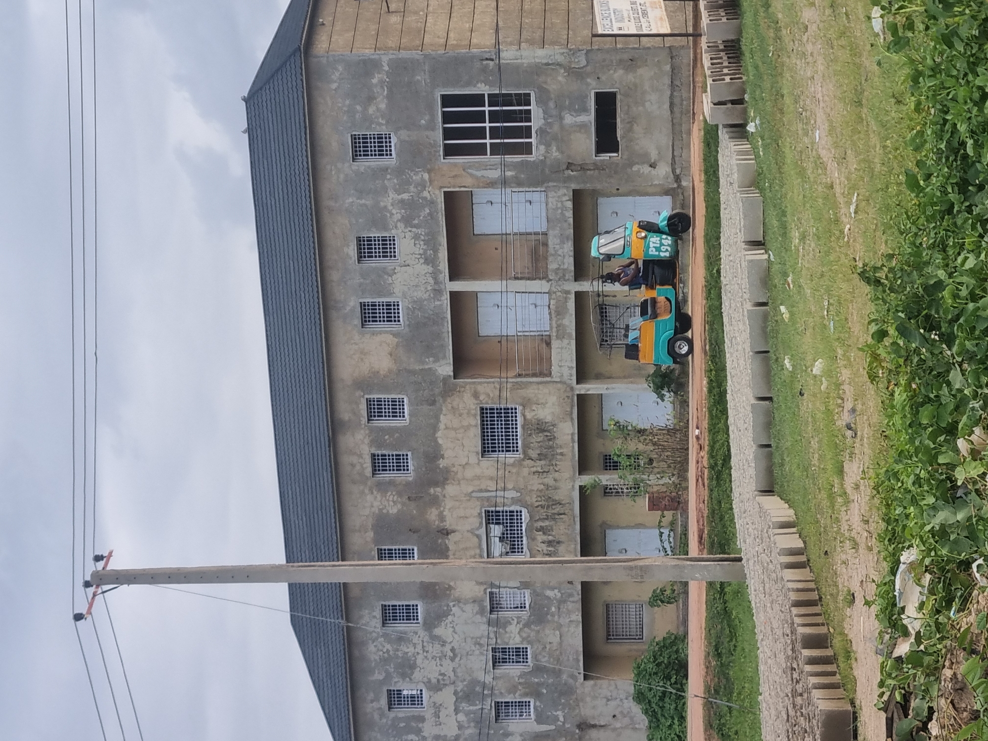 The image depicts a building in Gwong, Nigeria. The building is a two-story structure with a weathered, concrete facade and a dark grey, tiled roof. It features multiple windows with white bars on the upper floor and doors on the lower floor. To the left of the building stands a tall, grey utility pole with electrical wires strung across the sky. In front of the building, a man is operating a three-wheeled vehicle, a tuk-tuk, which is painted in blue and yellow and has the text "PTA 1943" on its side. The foreground shows rows of concrete blocks arranged on a grassy area, suggesting a construction or building materials site. The sky is overcast with clouds. A signpost is visible on the right side of the building, partially obscured.