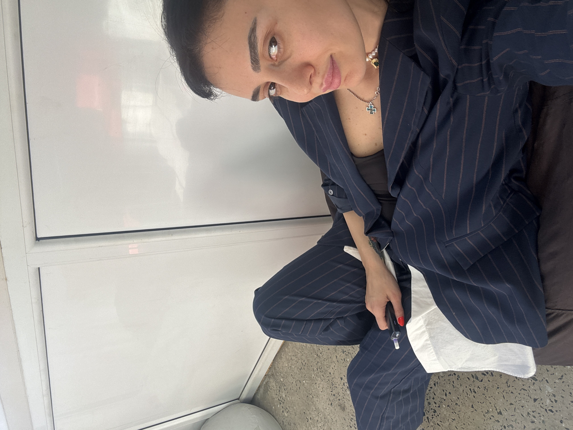 A woman is featured in a high-angle, rotated selfie, captured indoors. She gazes directly at the camera with a calm, slightly smiling expression. Her dark hair is styled back, and she has defined eyebrows and light brown eyes. She is dressed in a chic navy blue pinstripe suit, comprising a blazer and matching trousers, worn over a dark top. Adorning her neck are two necklaces: one featuring a silver cross pendant and another with pearls and a green gem. Her fingernails are painted red, and she holds a dark, slender object, possibly a vape pen or stylus, in her right hand.
The background consists of plain white wall panels or doors with dark trim, indicative of a modern interior space. The floor beneath her is a light grey, speckled surface, resembling concrete. A piece of white fabric is visible near her lap, possibly part of her clothing or an accessory. A partial view of a light grey, rounded object rests on the floor to her left. The lighting is bright and evenly distributed throughout the scene. This image depicts a moment in Tbilisi, Georgia.