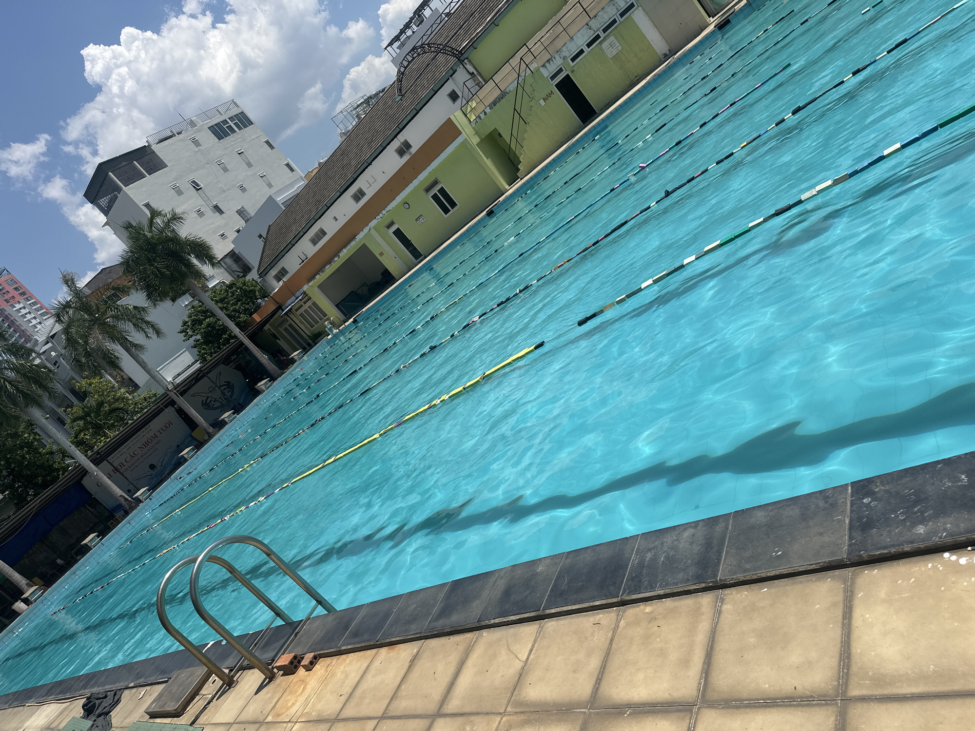 The image captures a swimming pool in the city of Nha Trang Ward, Vietnam. The pool, filled with clear turquoise water, has multiple lanes marked by yellow and black lane lines. A stainless steel ladder provides access to the pool. The pool's edge is defined by dark gray tiles, which border tan-colored square tiles that extend outwards. In the background, there is a mix of buildings, including a white high-rise with multiple windows, a green and white building with exterior walkways and windows, and a partially visible pink high-rise. Palm trees are scattered in front of the buildings. The sky is a light blue with scattered white clouds, suggesting a sunny day. A sign with Vietnamese text is also visible to the left of the frame.