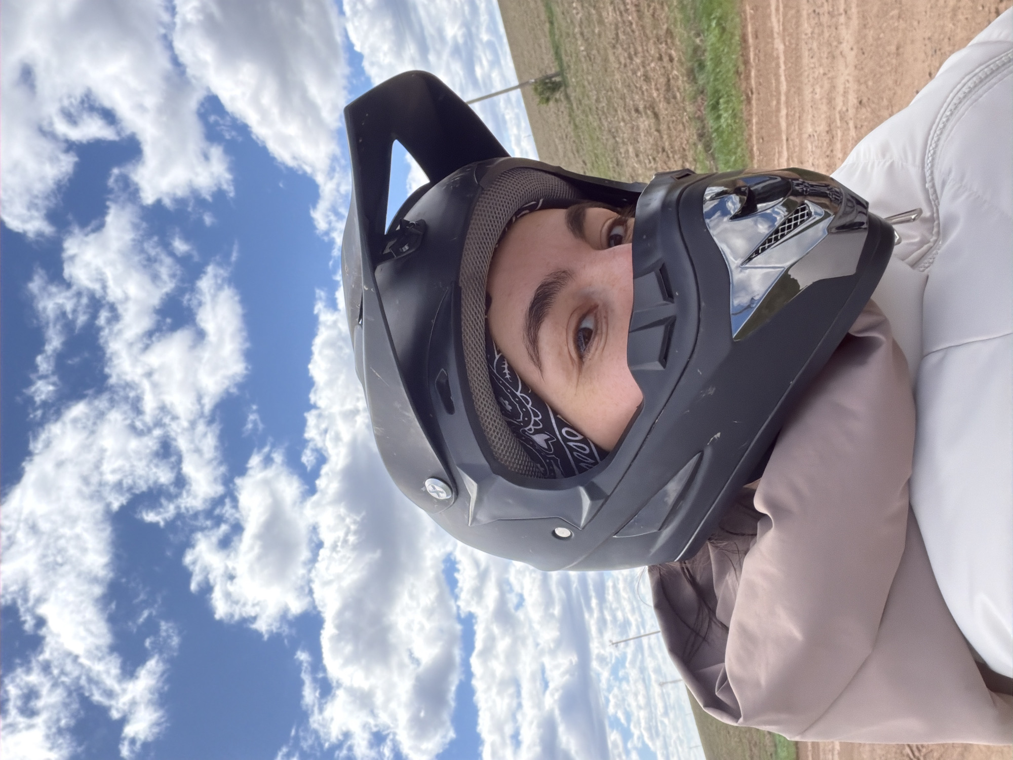 A person wearing a black motocross helmet is seen in a close-up, angled view. The helmet has a visor and a peak, and a bandana is visible around the person's neck inside the helmet. The person's eye is visible. The background is a field and a partly cloudy blue sky. The location cannot be determined to be Mozhga, Russia from the image alone.