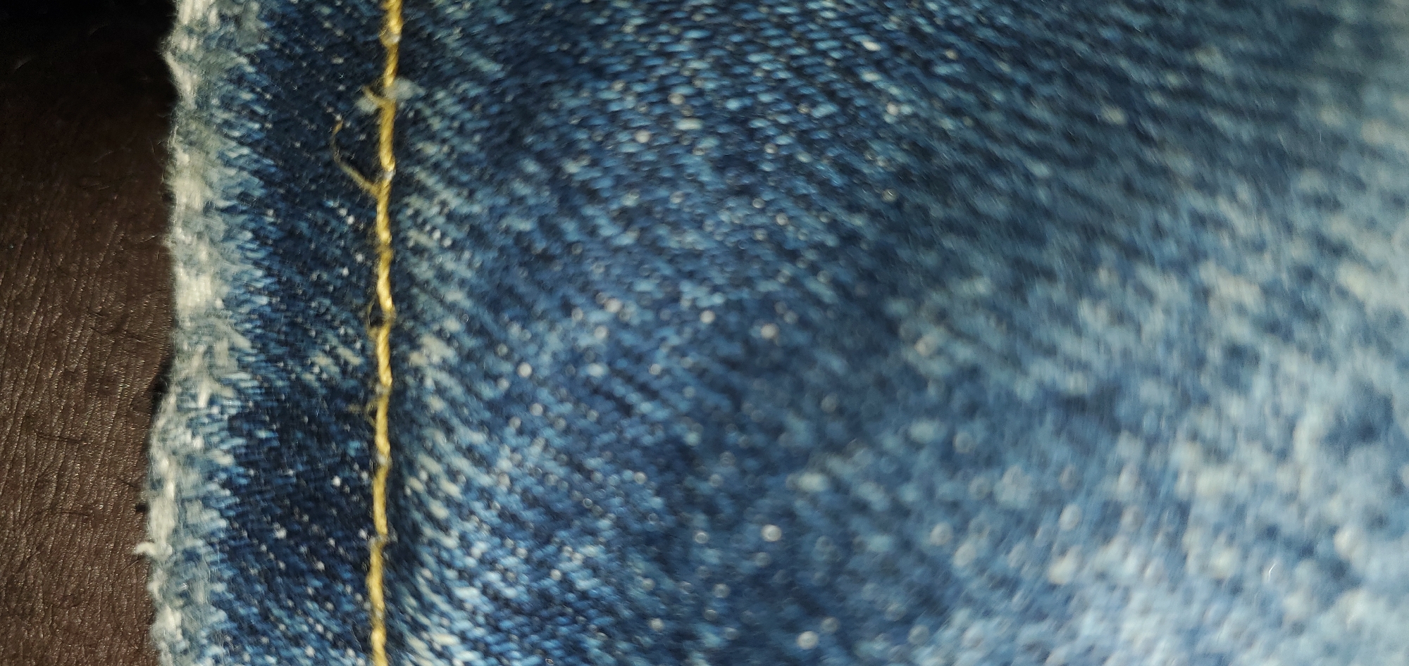 A close-up, slightly out-of-focus shot shows the texture of blue denim fabric. A single strand of gold thread runs horizontally across the top third of the frame. The denim appears slightly worn, with lighter speckles and a frayed edge visible at the very top. Above the denim, a dark, textured surface, possibly leather or a similar material, is visible. The image is devoid of any contextual clues regarding location or specific setting within a city.