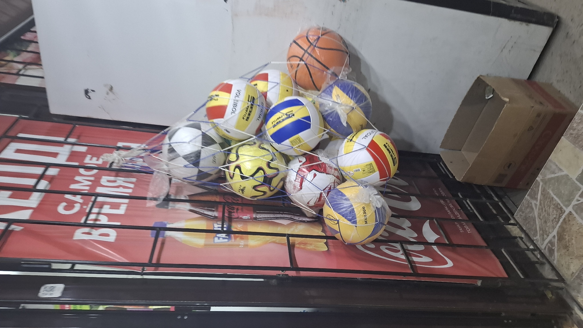 The image depicts a street-level shop display in Shymkent, Kazakhstan. A metal grate encloses the front of the shop, behind which several sports balls are displayed in a net. The balls include soccer balls, volleyballs, and a basketball. A large red banner with white text is visible behind the balls, featuring Russian text. Two bottles of a beverage are also visible behind the grate. To the right of the display, a plain white wall and a cardboard box are present. The ground in front of the shop is tiled. The scene suggests a retail environment, likely selling sporting goods or related merchandise.