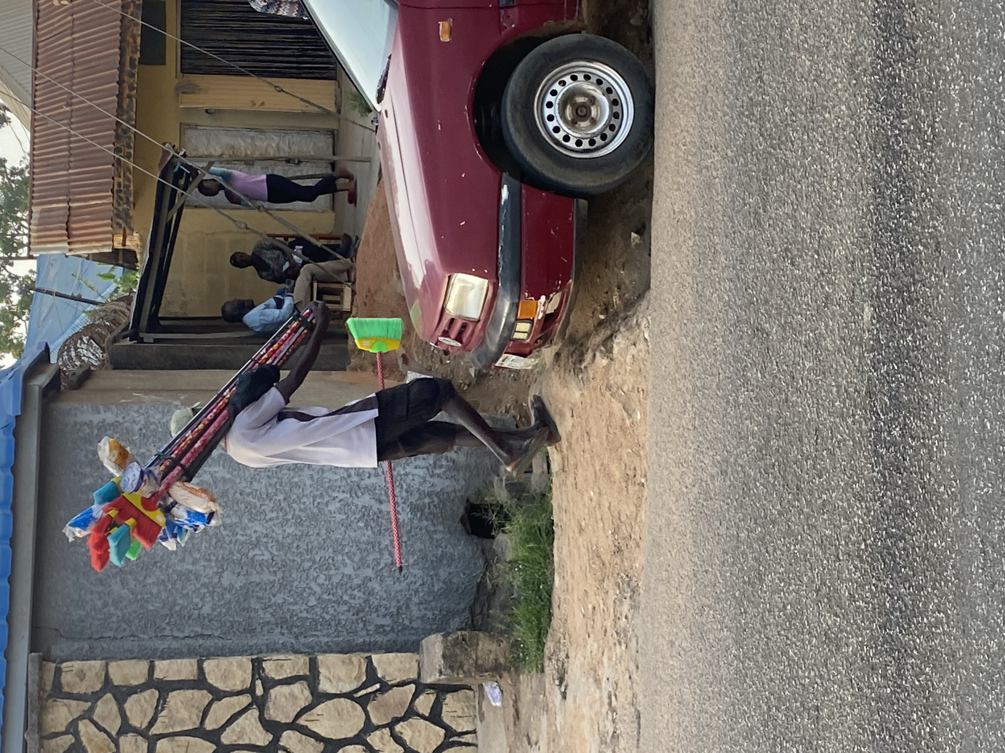 The image depicts a street scene in Jos, Nigeria. A man is walking along the side of a road, carrying a collection of colorful, plastic brooms and brushes for sale on a stick over his shoulder. He is wearing a white top and dark shorts. The foreground includes a section of asphalt road on the right and a roadside area with exposed dirt and some green vegetation.
A parked, red car partially occupies the upper right of the frame. Behind the man and to the left, there is a stone wall and a blue-roofed structure. Further in the background, across from the car, are people sitting and reclining in front of a building. The setting appears to be an urban or semi-urban environment with everyday activity.