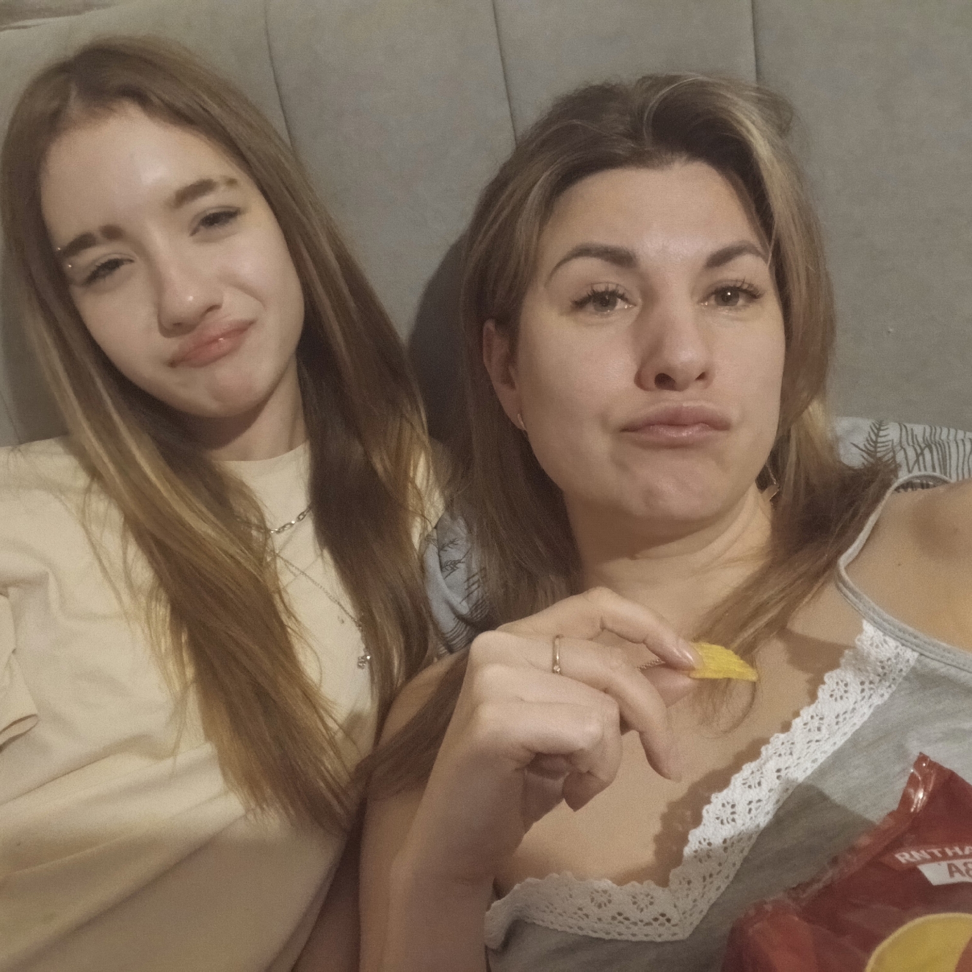 Two women are lying down, likely in a domestic setting. The woman on the left has long brown hair, a light-colored shirt, and an eyebrow piercing. The woman on the right has shoulder-length brown hair, a grey tank top with lace trim, is holding a potato chip, and has a bag of chips nearby. The background appears to be a grey upholstered headboard. This is unlikely to be Izhevsk, Russia as it is not easily discernible from the image.