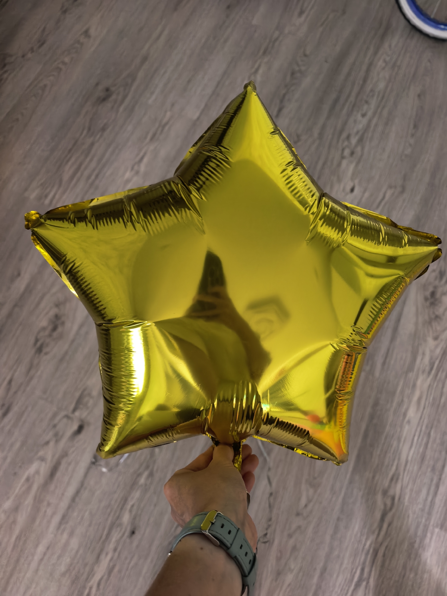 A human hand, appearing to be an adult's right hand, holds a fully inflated, metallic gold, five-pointed star-shaped Mylar balloon. The hand is positioned centrally at the bottom of the frame, gripping the balloon at its sealed neck. A grey or mint green watch or wristband with a yellow buckle is visible on the wrist. The balloon exhibits strong reflections of its surroundings, including what appears to be a distorted reflection of the person holding it. The background consists of a light-toned wooden floor with planks laid diagonally, running from the lower left to the upper right. In the upper right corner of the frame, a segment of a white bicycle tire with a blue rim is visible. The scene depicts an indoor environment.