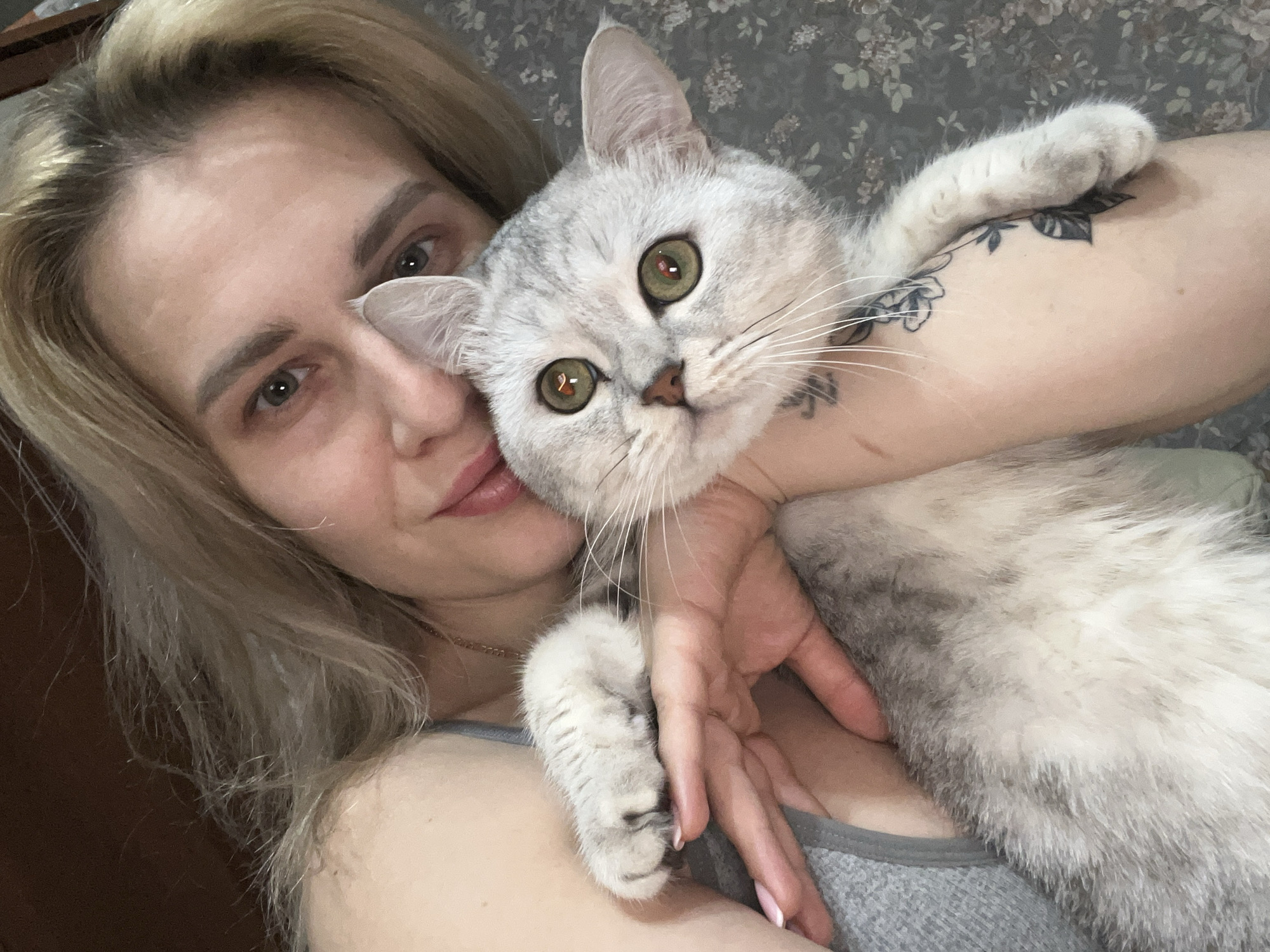 A fair-skinned woman with blonde hair is holding a gray and white cat. The woman's left arm is around the cat's neck. The cat's front left paw is in the woman's hand, and the cat's front right paw is near the woman's right arm, where there is a tattoo. The woman is wearing a gray tank top and a necklace. The background appears to be a gray floral patterned fabric. The location is in Chaikovsky, Russia.