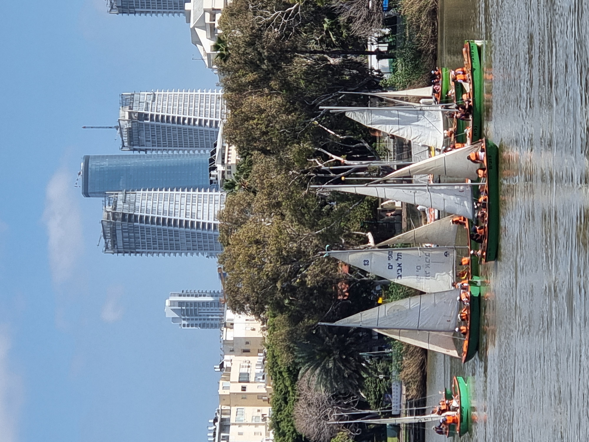 Several green sailboats with white sails are on a body of water, likely the Yarkon River, in Tel Aviv, Israel. Each boat contains several people wearing orange life vests. The boats are arranged in a line, moving in the same direction. The background consists of a skyline of Tel Aviv buildings, trees, and other urban greenery. The sky is blue with scattered clouds.