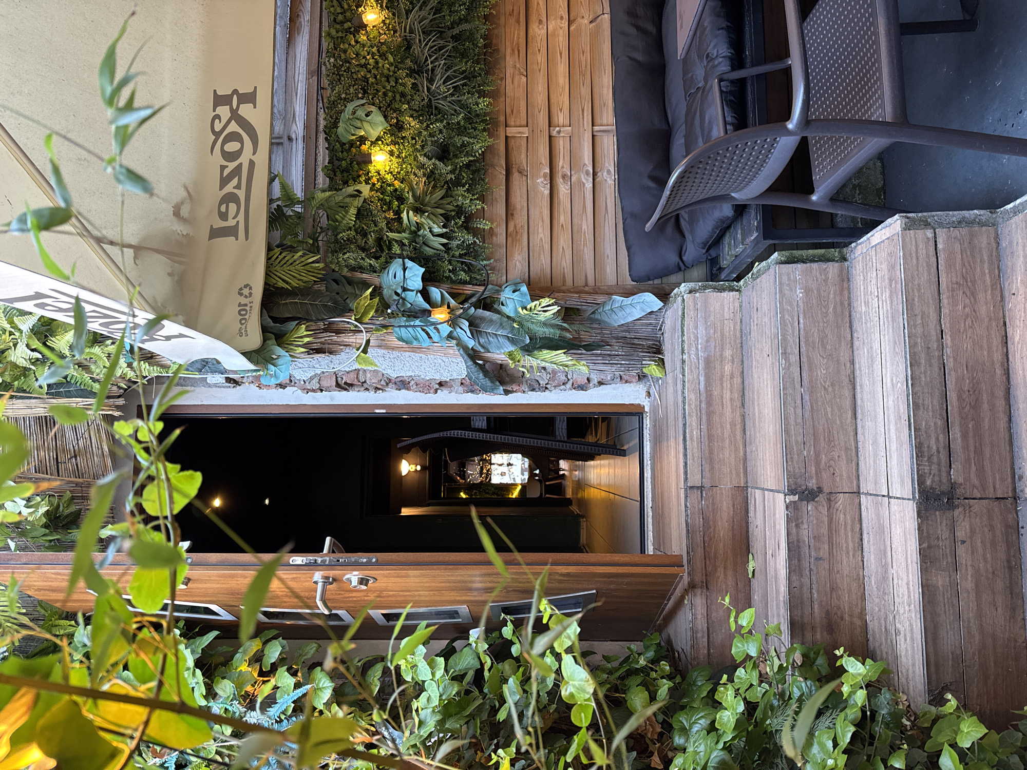 The image displays the entrance and a proximate outdoor seating area of an establishment. A set of dark wooden stairs, consisting of approximately six risers, ascends from the foreground towards an open, single wooden door. Dense green foliage, resembling ivy, covers the ground and a low wall to the left of the staircase. The wooden door is equipped with silver hardware and reveals a dimly lit interior hallway with tiled flooring, warm light fixtures, and a partially visible dark chair.
To the right of the entrance, an outdoor seating zone features a retracted beige awning, which displays the inverted text "Kozel" and a "100% rec..." logo. Below the awning, a vertical garden wall, composed of various green plants including ferns and larger-leafed species, is illuminated by three warm-toned filament light bulbs. This green wall is situated above a horizontally slatted wooden panel. In front of the panel, a black bench with a thick black cushion is present. A dark brown, perforated plastic chair with armrests occupies the right foreground. Sections of exposed brickwork are visible on the far-right wall, and woven bamboo-like material is present above the door and on the far-left wall of the outdoor area. The scene is illuminated by a combination of ambient light and warm artificial sources.