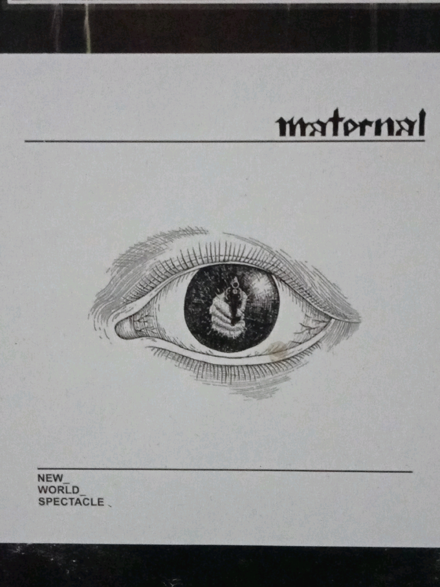 The image is a black and white drawing of a human eye. The iris of the eye contains a reflection of a pistol. Above the eye, the word "maternal" is printed in a gothic-style font. Below the eye, the words "NEW WORLD SPECTACLE" are printed. The background is plain white. There is no identifiable location in Depok, Indonesia.