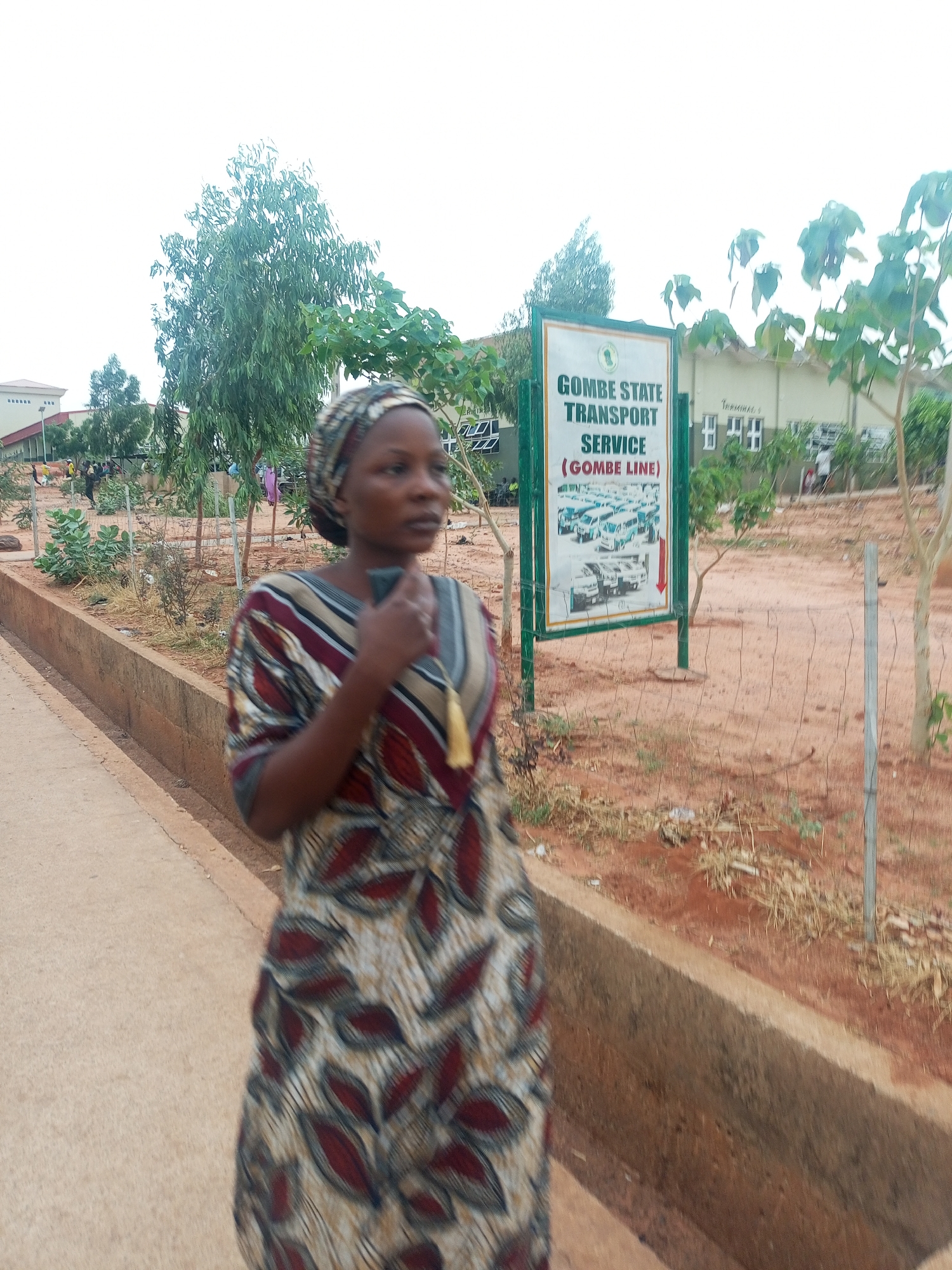 A woman wearing a patterned dress and headscarf is walking near a sign that reads "Gombe State Transport Service (Gombe Line)." The sign has an image of vehicles. There are trees, buildings, and a sidewalk in the background. The location is Gombe, Gombe State, Nigeria.