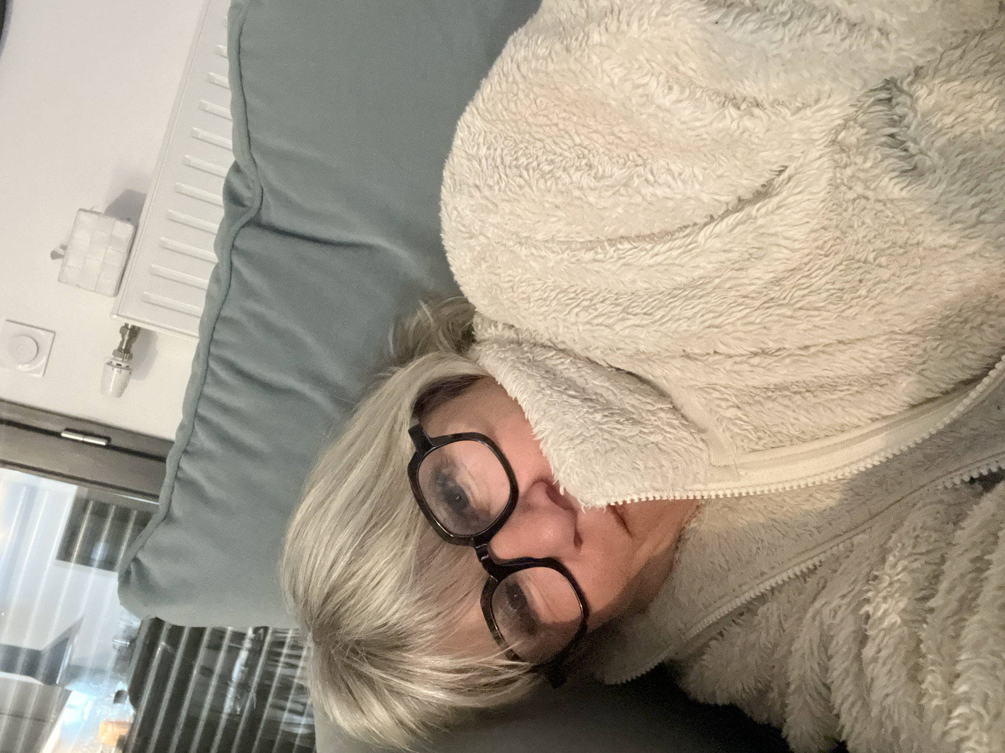 A woman lies on a light blue pillow, partially covered by a white fuzzy jacket, indoors. She wears black-rimmed glasses. The room features a white wall with a radiator and valve. The location is Chambœuf, France.