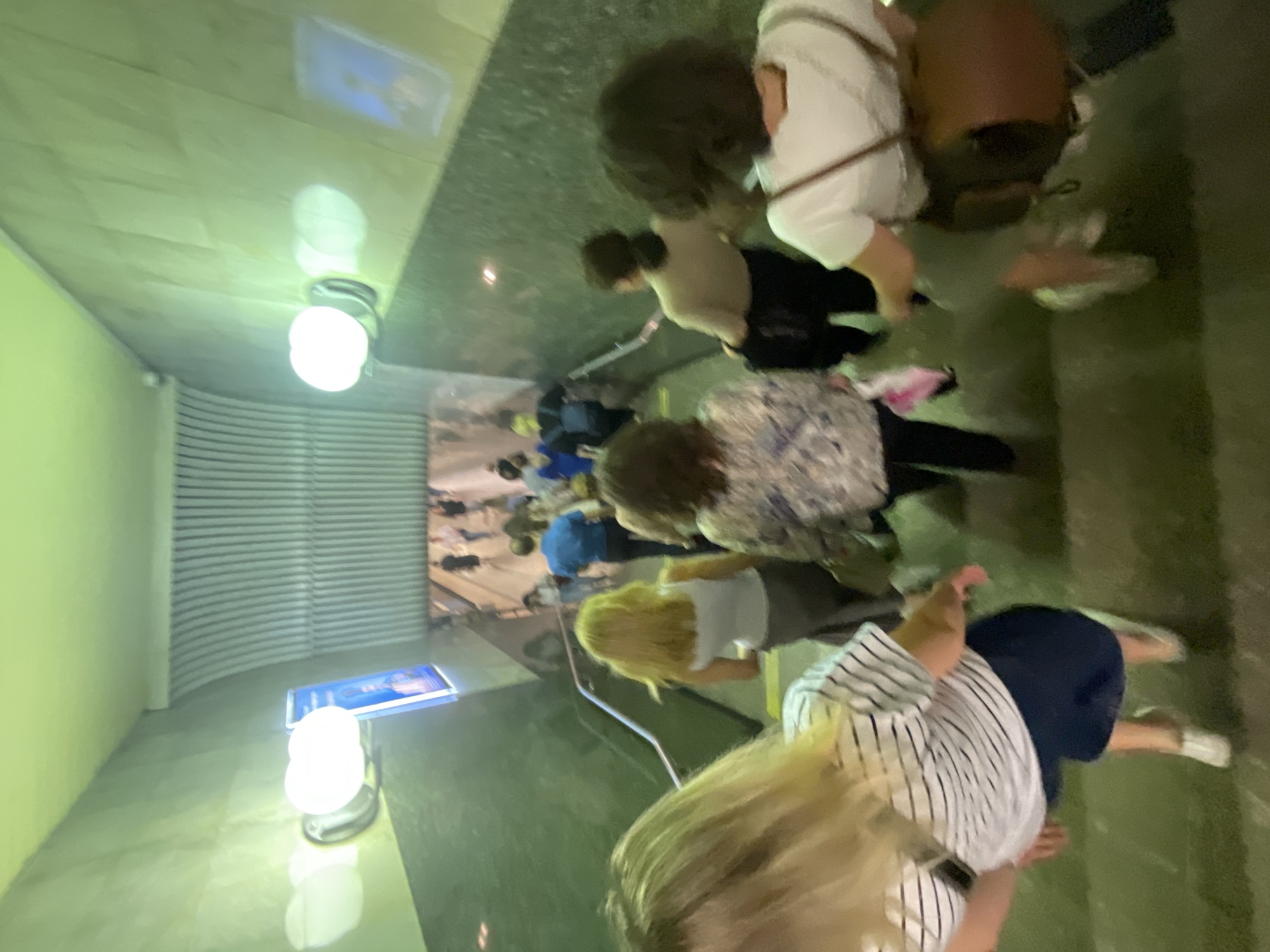 The image shows a group of people walking down a staircase in a subway station. The station appears to be underground, with green tiled walls and fluorescent lighting. In the foreground, a woman with blonde hair wearing a striped shirt and dark blue skirt is walking down the stairs. Behind her, a woman with dark brown hair wearing a patterned shirt and black pants is also descending. Further down the stairs, more people are visible, including a man in a blue shirt and a woman with blonde hair in a white top. The overall impression is of a busy urban environment, likely during a commute. The specific location in Moscow, Russia, cannot be determined from the image alone.