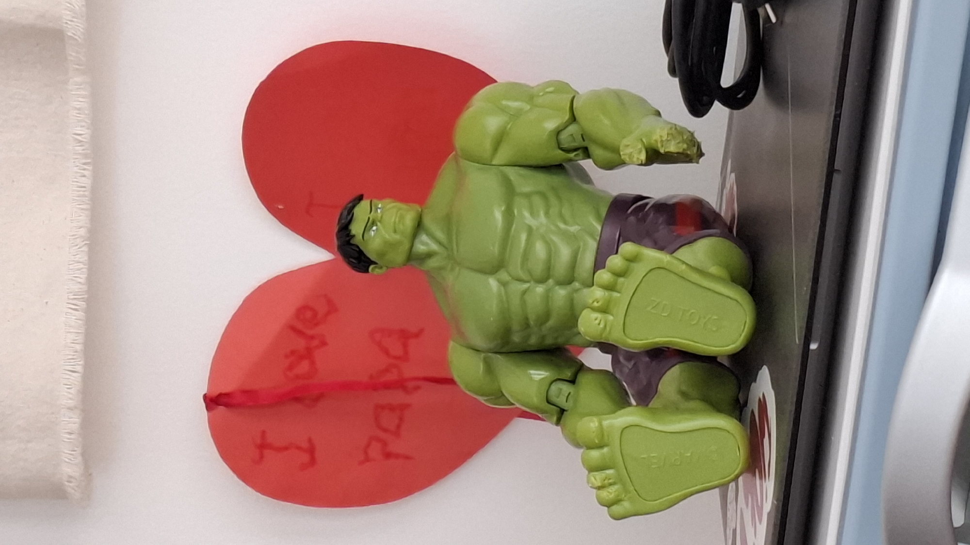 The image shows a Hulk action figure lying on its side, partially on a heart-shaped red paper card with the handwritten message "I love Papa." The Hulk figure is green with purple shorts, and the words "ZD TOYS" and "MARVEL" are visible on its feet. The figure and card are on top of what appears to be a crafting machine or other electronic device. A white cloth is visible on the left side of the frame, and various cords and parts of the machine are visible on the right. There is no information indicating location or city.