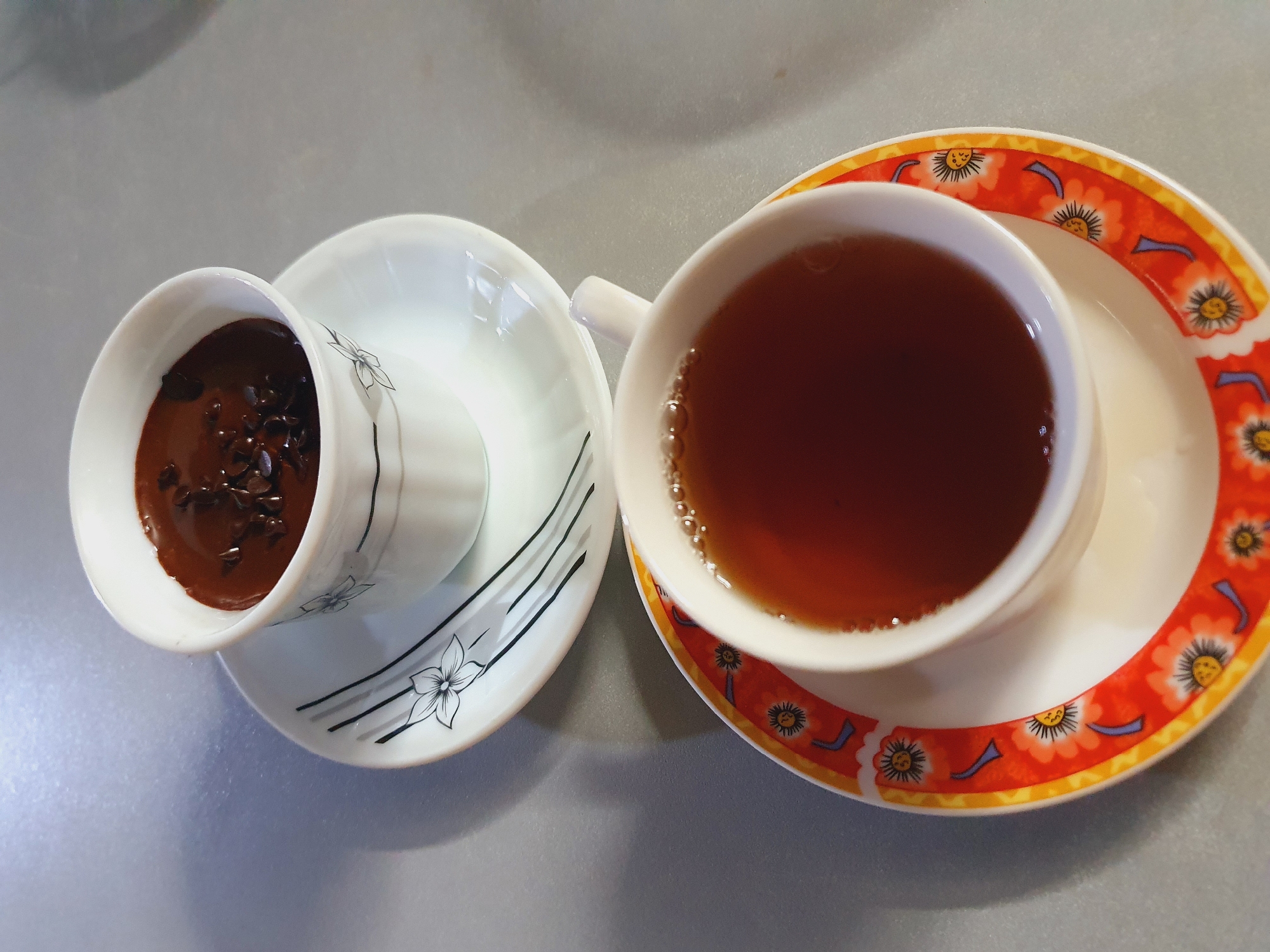 This image, taken at a residence in Shiraz, Iran, features two coffee cups on saucers arranged on a gray, reflective surface. The upper cup, smaller in size, contains a dark brown liquid, possibly hot chocolate, topped with chocolate shavings. This cup has a white exterior with delicate black floral designs and lines. The saucer beneath it is plain white. The lower cup is larger and filled with a reddish-brown liquid, likely tea, with small bubbles visible on the surface. Its saucer is white and adorned with a vibrant orange border featuring floral patterns in yellow and blue. The lighting suggests an indoor setting, possibly a kitchen or dining area, with soft shadows cast on the surface.