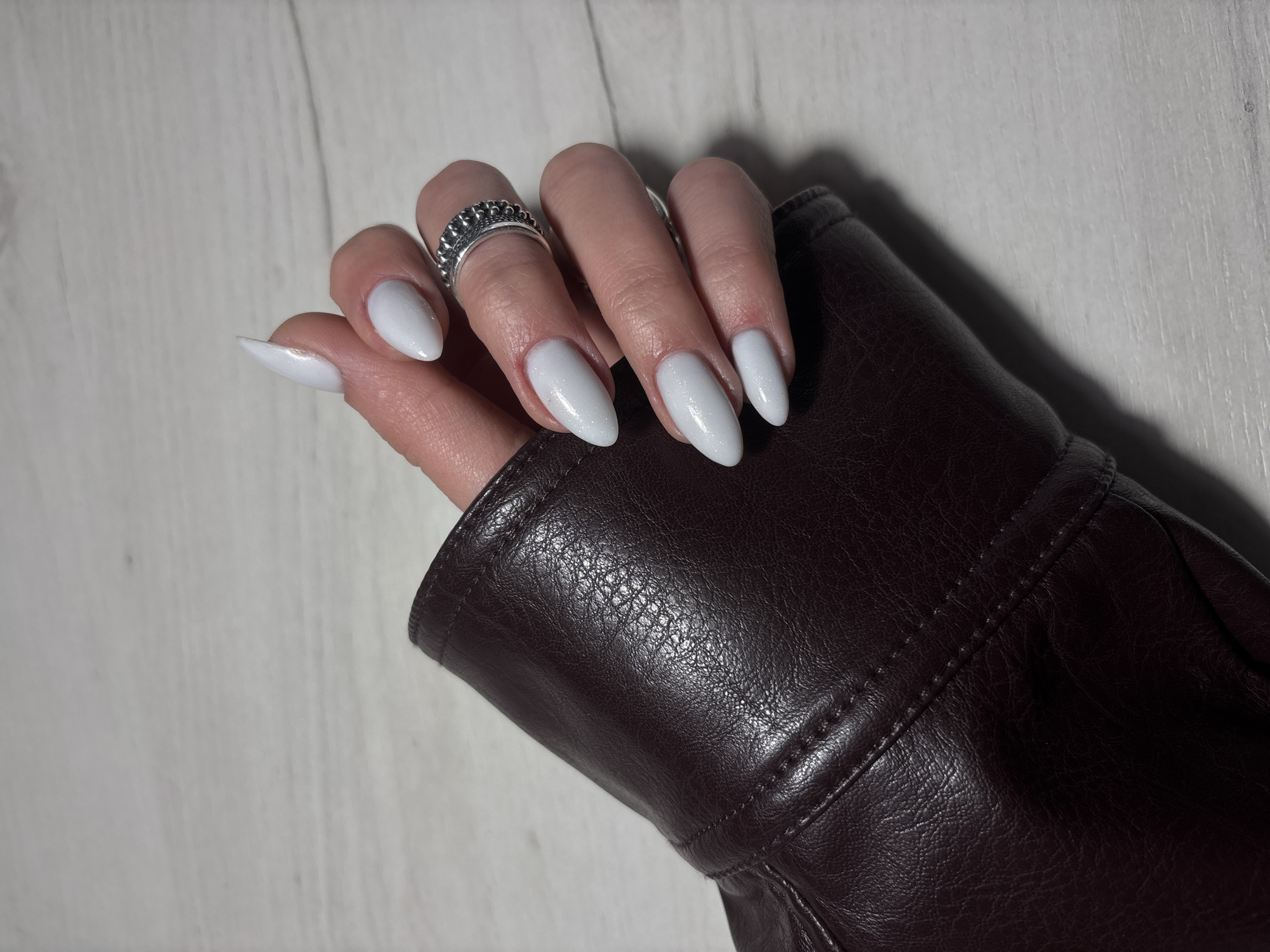 A person's hand, adorned with a silver ring and painted nails, rests on a dark brown leather sleeve. The fingernails are long, almond-shaped, and painted with a light, possibly white or off-white, color. A silver ring with a textured design is worn on the ring finger. The dark brown leather sleeve is visibly textured. The hand and sleeve rest on a light-colored wooden surface. There is no indication of the location being Yerevan, Armenia in the provided image.