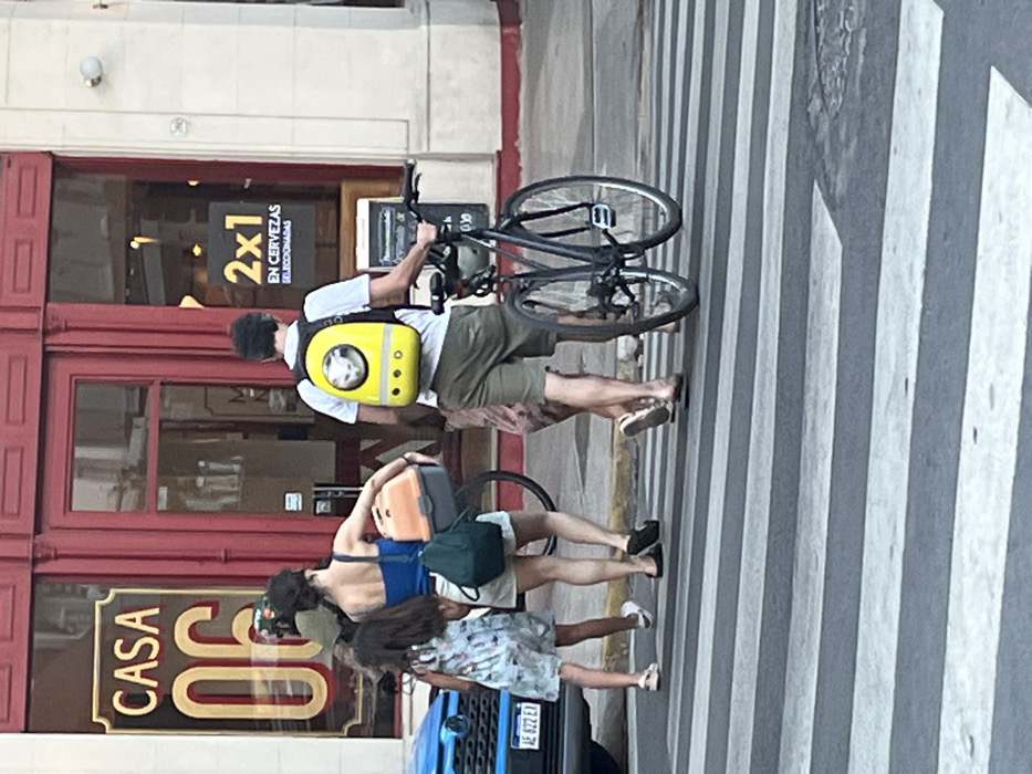 A group of people walks across a crosswalk. One person carries a bicycle over their shoulder and wears a bright yellow backpack. Another person carries two bags, while a third person walks alongside them. A building with a sign that reads "CASA 06" is visible in the background. The door of the building is red and has a sign that says "2x1 EN CERVEZAS SELECCIONADAS". A car is parked to the left of the group.
