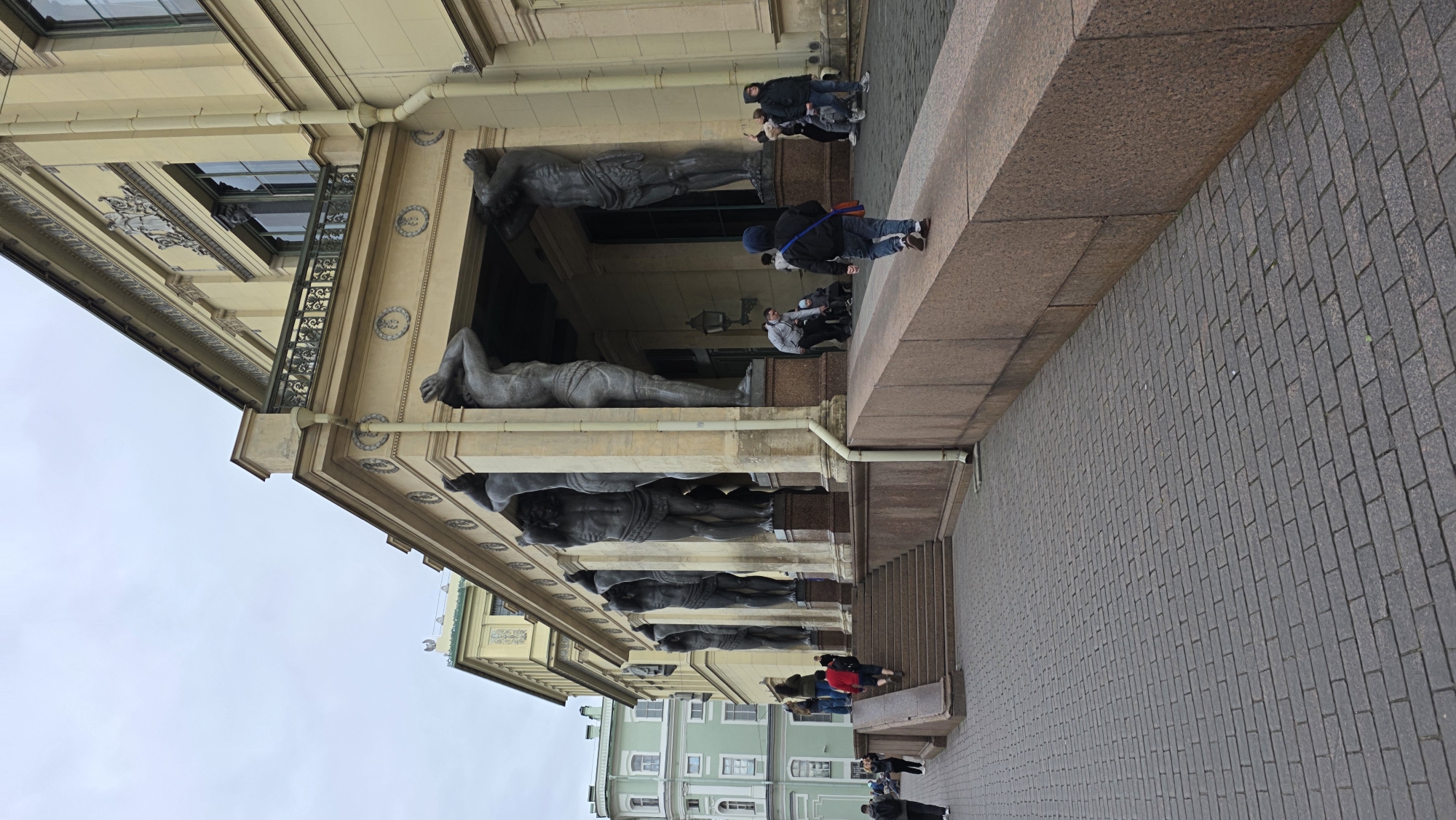 Here is a detailed description of the image:
The image is taken from a low angle, capturing a section of an imposing building with architectural features resembling classical design. The building's facade has a series of colossal statues of men supporting the structure above. These statues are gray in color and have a muscular build.
Several people are seen both inside and around the building. Some are standing on the pavement, while others are near the building's entrance, observing or taking pictures. The pavement is made of small, rectangular blocks.
The building has ornate decorative elements such as patterns, moldings, and a balcony with iron railings. The sky is overcast, suggesting a cloudy or rainy day. The building's color palette consists of light yellows and grays.
Based on the architectural style and the presence of the large statues, this location is likely the portico of the New Hermitage building in Saint Petersburg, Russia, renowned for its distinctive row of Atlantes statues.