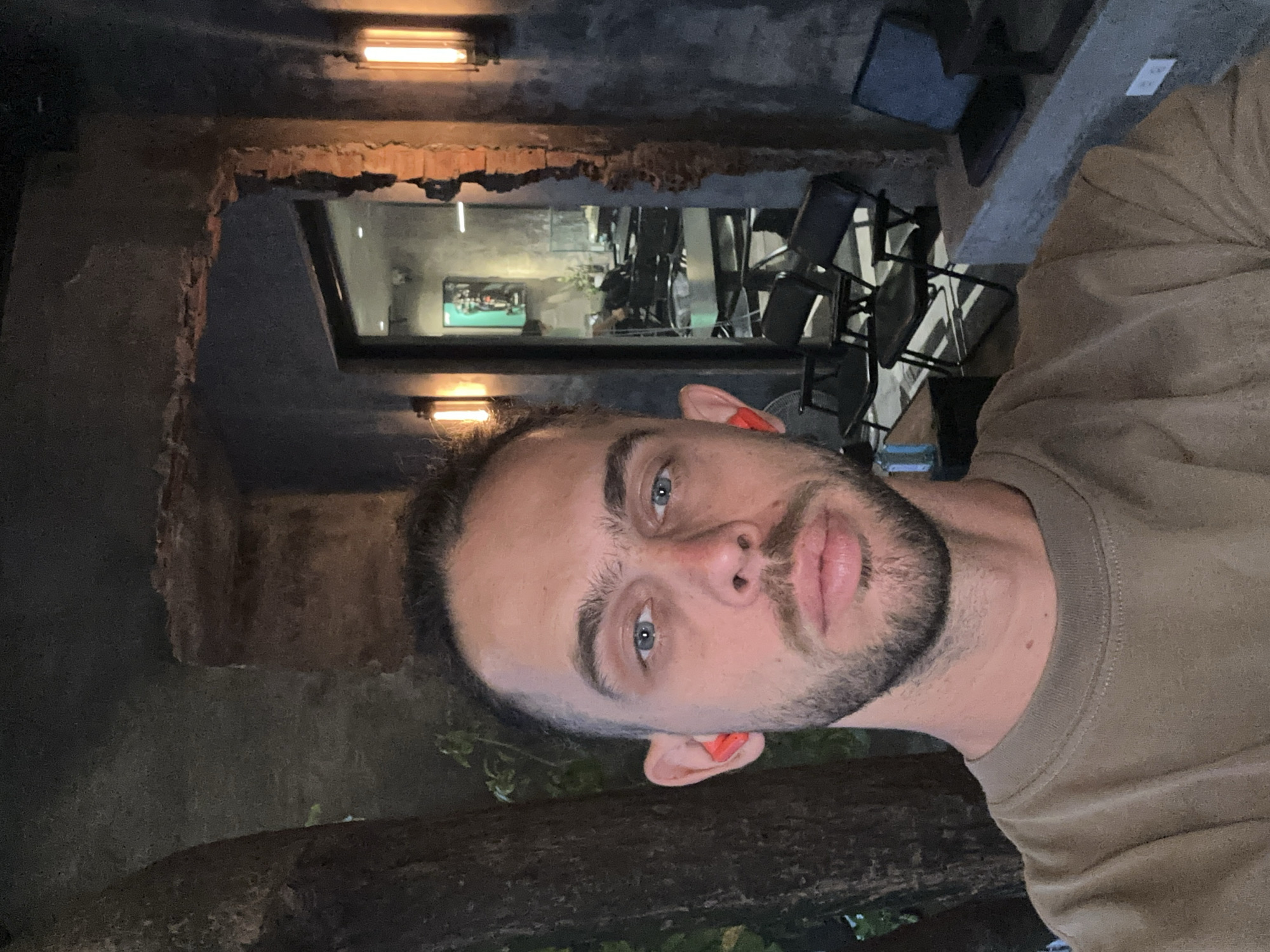 A man with blue eyes and a beard, wearing orange earplugs and a brown t-shirt, stands in front of a rustic, exposed brick archway. To his left is the trunk of a large tree. Beyond the archway, inside a dimly lit establishment, rows of black chairs are visible, suggesting a salon or waiting area. Two vertical wall sconces with warm lighting are mounted on the exterior wall. The scene appears to be outdoors in Đà Nẵng, Vietnam, judging by the architectural style and vegetation.