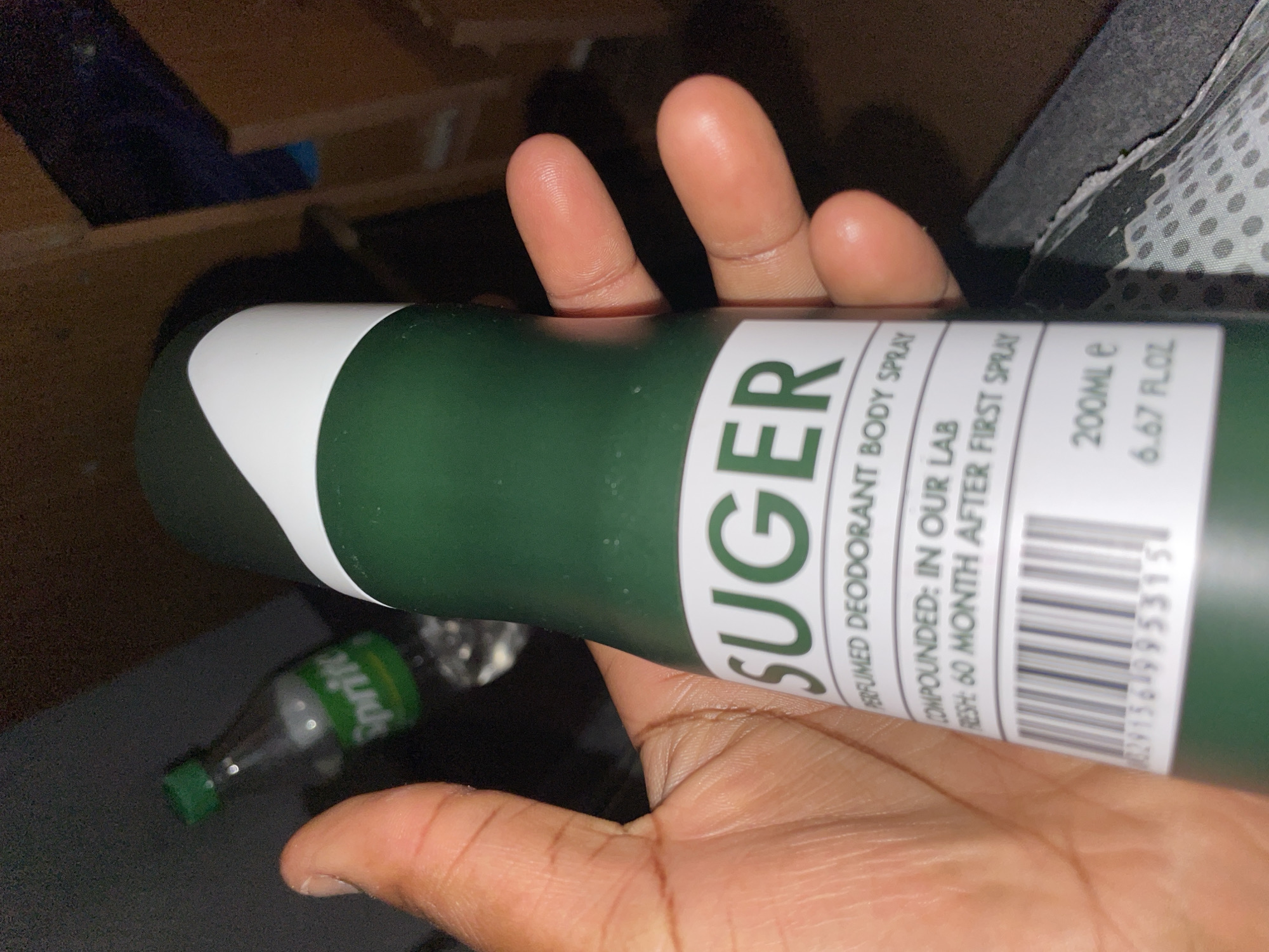 A person is holding a green can of SUGER perfumed deodorant body spray in their hand. The can has a white top. The product details visible on the can include "COMPOUNDED: IN OUR LAB", "FRESH: 60 MONTH AFTER FIRST SPRAY", "200ML e", and "6.67 FLOZ", along with a barcode and corresponding number. A small bottle of Sprite is partially visible in the background on the left side of the image. Other objects are visible in the background, including some possibly made of wood. The location of Kurunduma, Nigeria, is not apparent in the image, and there is no indication that it was taken there.
