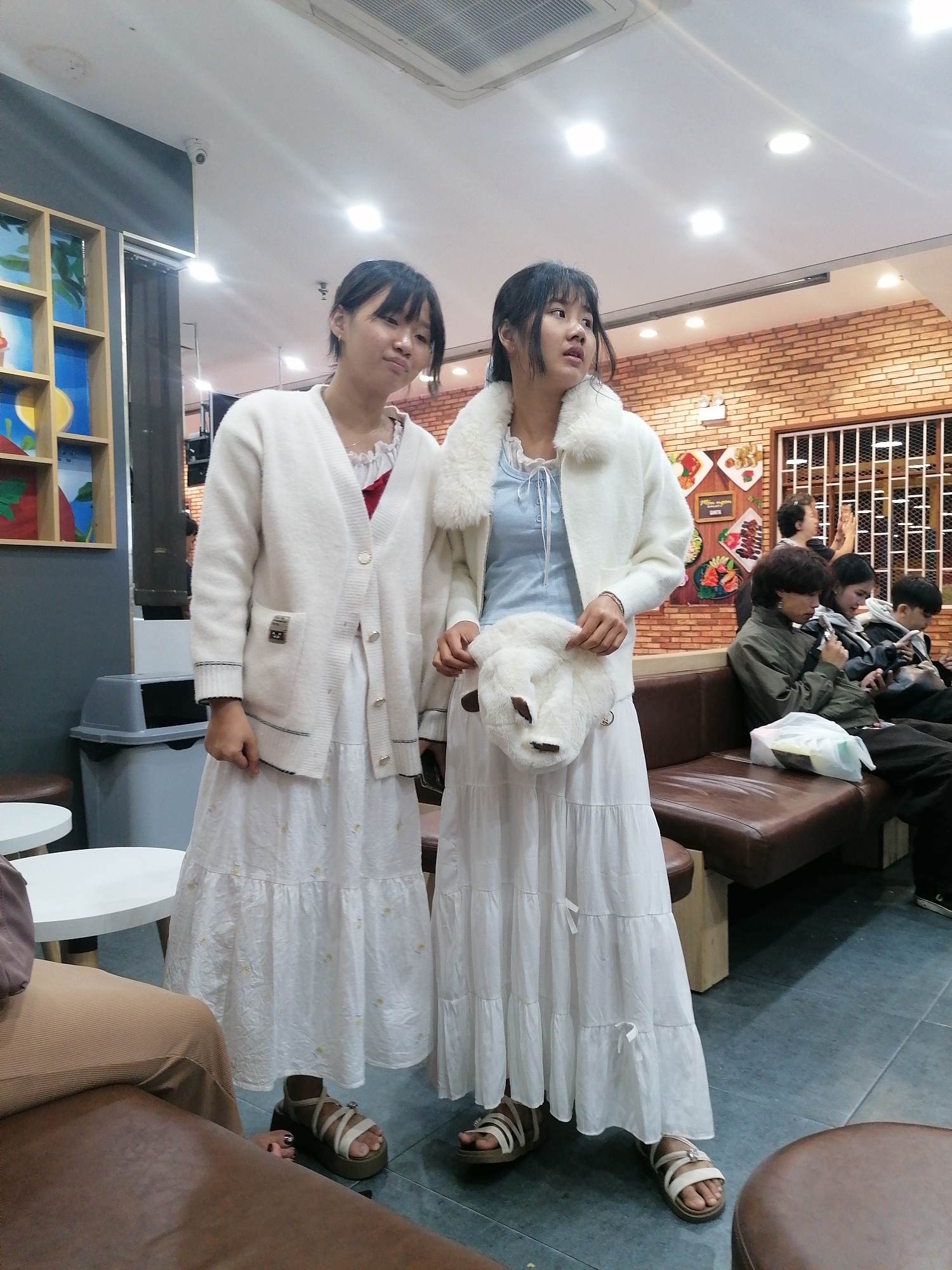 Two young women stand in a brightly lit indoor space that appears to be a cafe or restaurant. The woman on the left wears a white cardigan over a white ruffled collar dress and white sandals. She is looking slightly to her left with a neutral expression. The woman on the right wears a white fluffy-collared jacket over a light blue top and a tiered white skirt, paired with white sandals. She is looking to her right with an open-mouthed expression. She holds a fluffy white purse shaped like a sheep. In the background, a brown leather sofa is occupied by several people, some of whom are looking at their phones. A wall with exposed brick and a decorative grill is visible behind them. To the left, a shelf unit displays colorful items. A grey trash can is also visible. The lighting suggests it is daytime or evening, with artificial lights on. The presence of a decorative brick wall and specific menu items on the wall suggests the location is likely a cafe or restaurant in Da Lat, Vietnam, a city known for its European-inspired architecture and cafes.