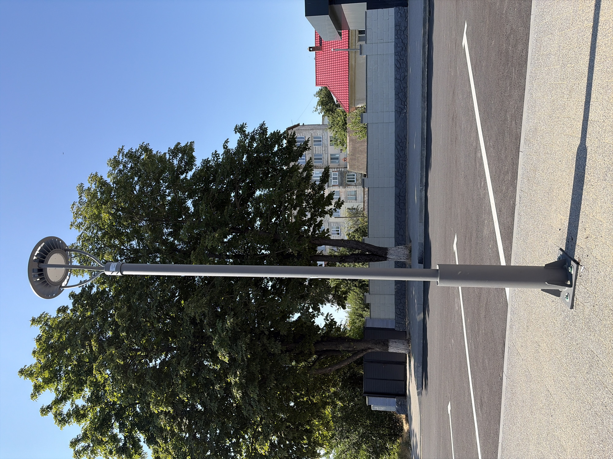 The image depicts an outdoor scene with a modern gray lamppost prominently featured in the foreground, situated on a light-colored, textured surface. Behind the lamppost, a paved parking lot is visible with white directional arrows painted on the asphalt. A low, gray stone and brick wall runs along the perimeter of the parking area, with a dark metal gate visible in the background to the left. Two large trees with full green foliage stand behind the wall. In the mid-ground, there are residential buildings, including a light-colored apartment building and a building with a red tile roof. The sky is clear and blue. The scene appears to be in the city of Criuleni, Moldova, based on the provided context. There are no people or animals visible in the image.