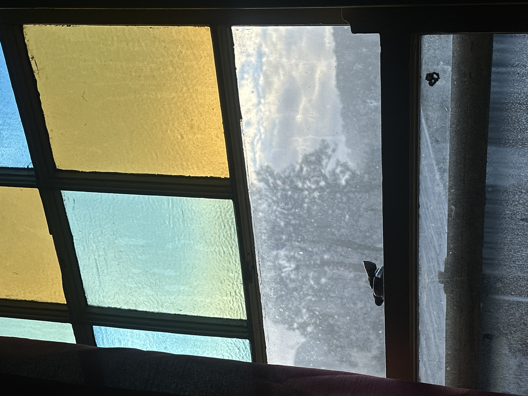 The image shows a window with stained glass in the upper portion and clear, frosted glass in the lower portion. The stained glass is divided into several panes, some of which are blue and yellow. There is a red curtain on the left side of the window. In the lower section of the window, a dark-colored latch mechanism is visible. The frosted glass obstructs the view of the outside, but some blurred shapes of trees and the sky can be discerned. The image is a close-up view and does not provide information about the location in Thora, Australia, nor does it contain any people or specific actions.