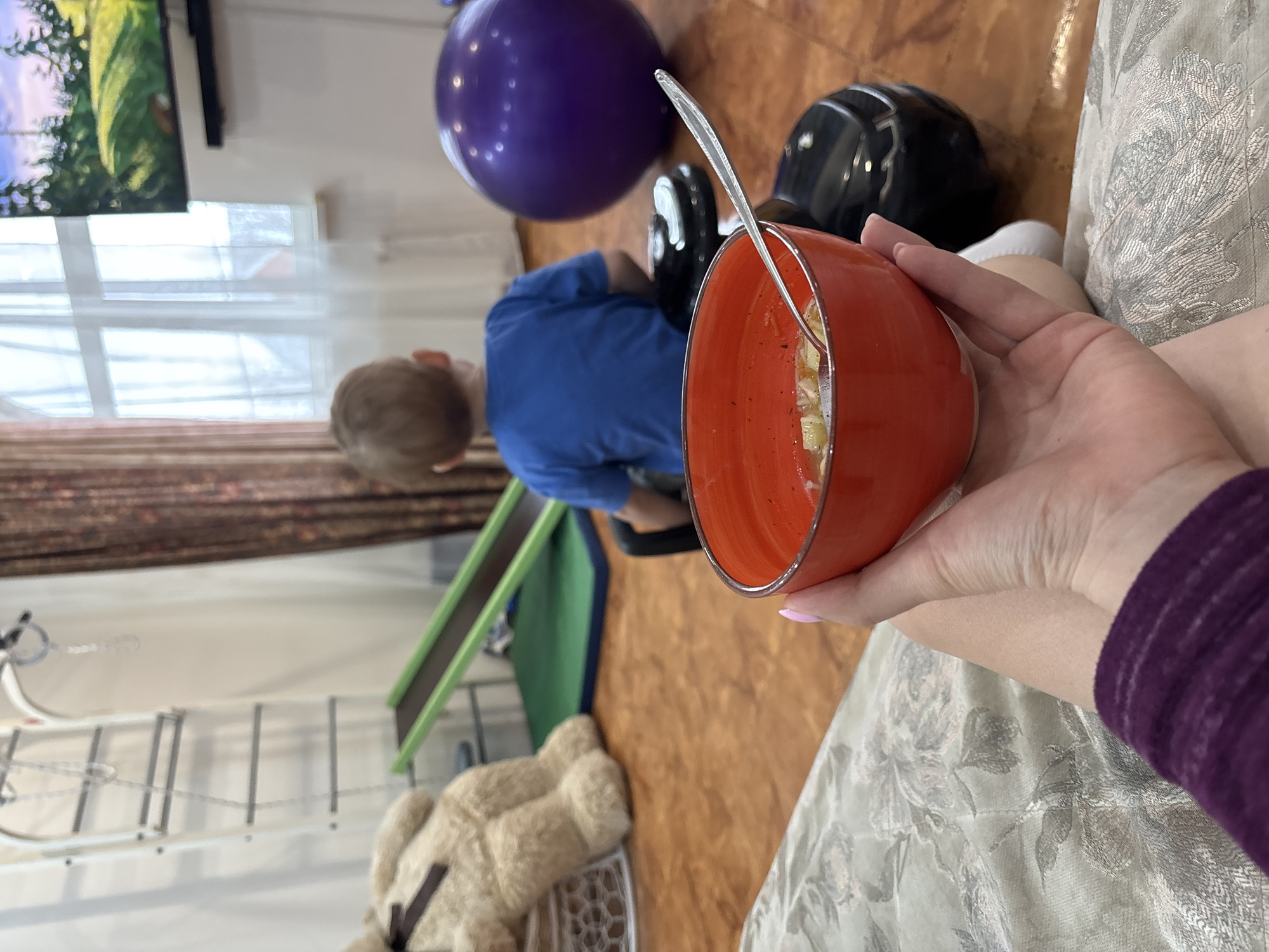 A person sits holding a red bowl of food with a silver spoon in their hand. A child wearing a blue shirt sits in the background, near a purple exercise ball and a black helmet. A children's slide, a teddy bear, and a set of climbing bars are also visible. The location is Ферма, Russia. A television is mounted on the wall, displaying a landscape scene.